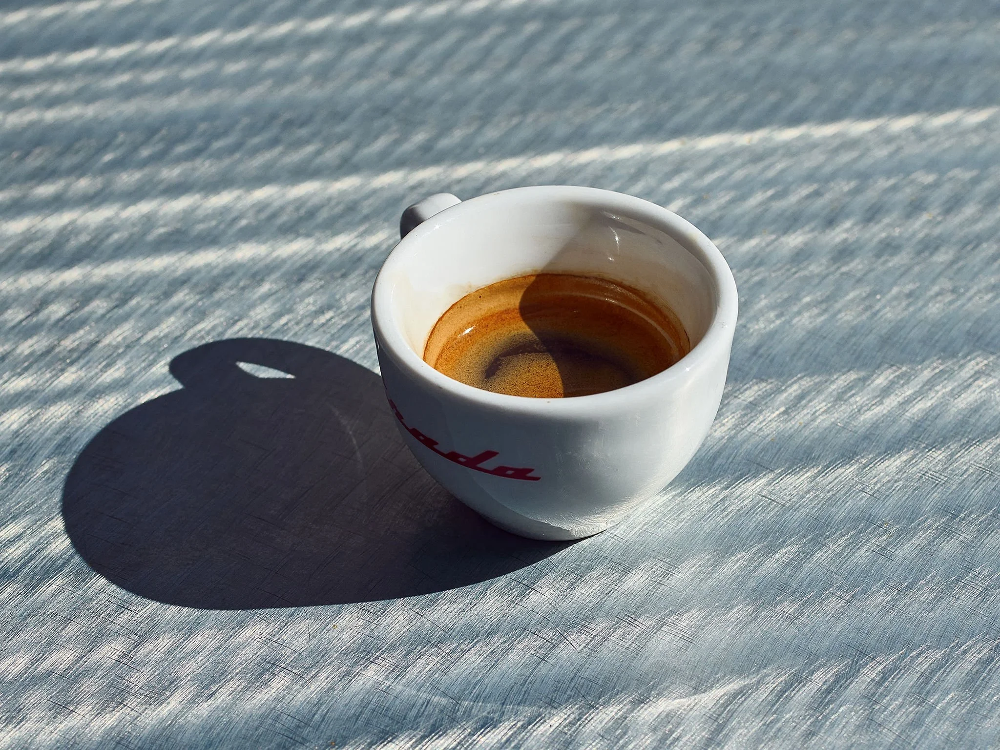 A small white espresso cup with red writing, filled with dark coffee, sitting on a silver textured surface with sunlight casting a shadow.