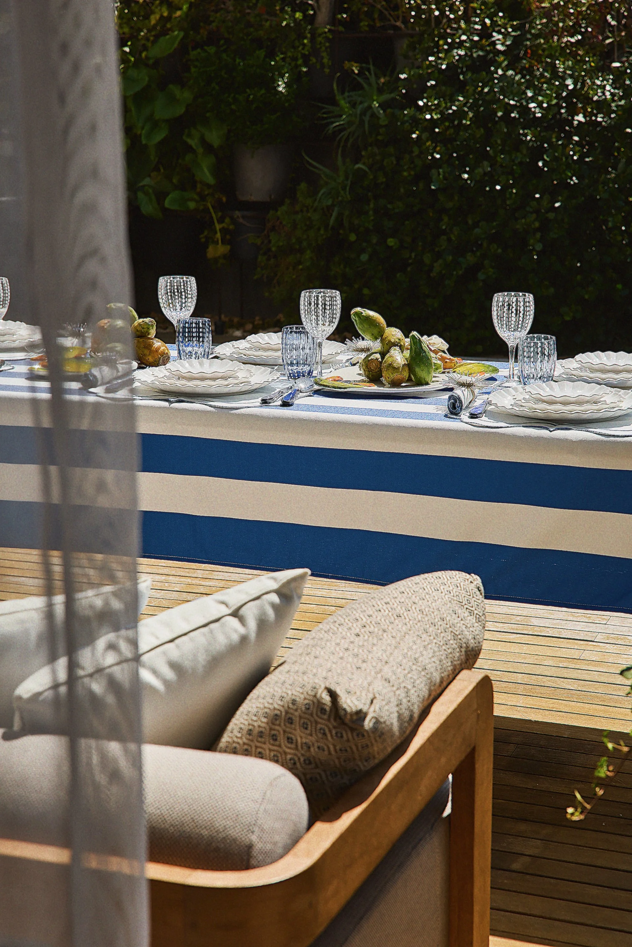 An outdoor dining table set with plates, glasses, and a platter of green vegetables. A couch with beige cushions and a patterned throw pillow is in the foreground, partially seen through a sheer curtain. The background features lush green foliage and