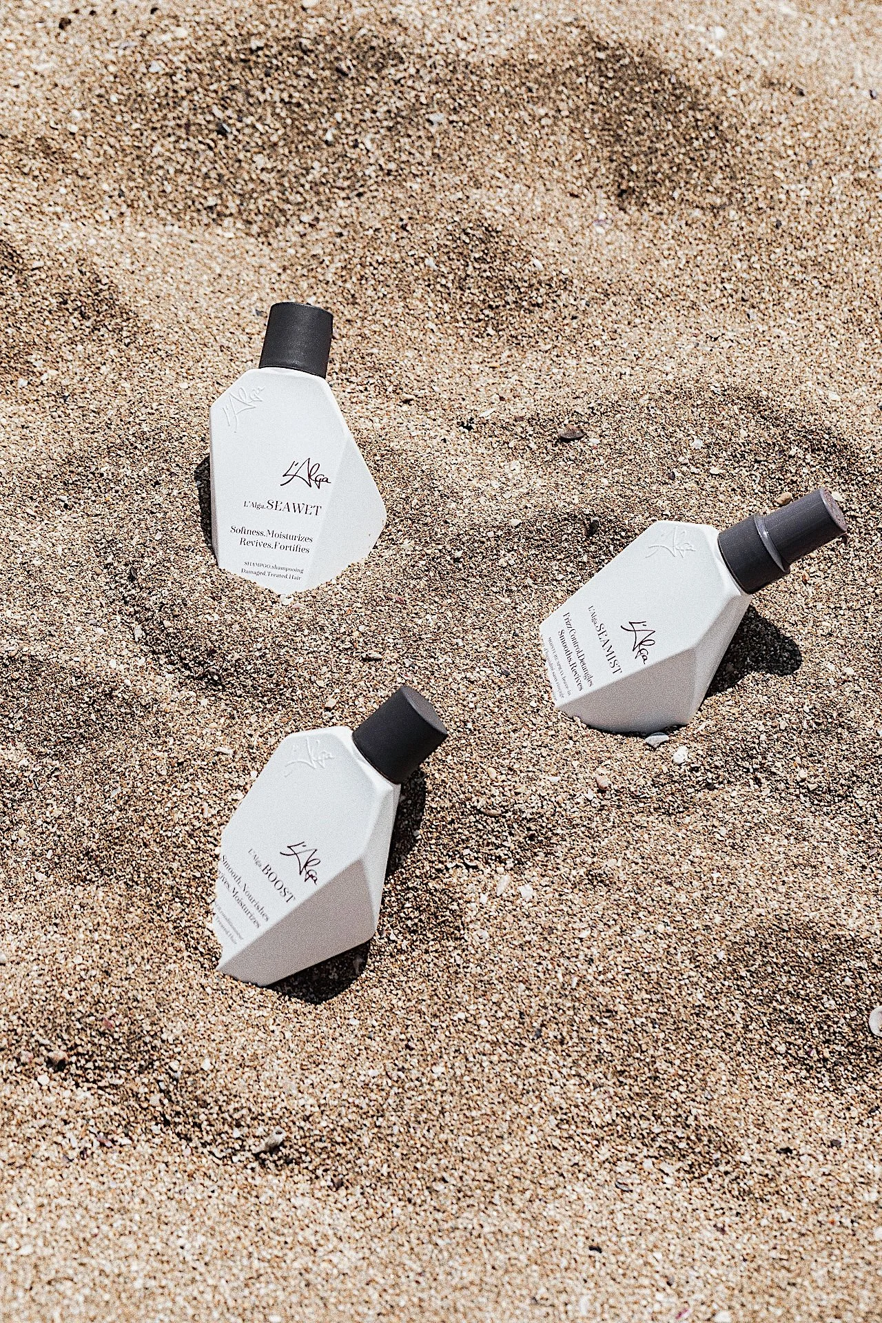 Three bottles of hair care products laying on sandy beach.