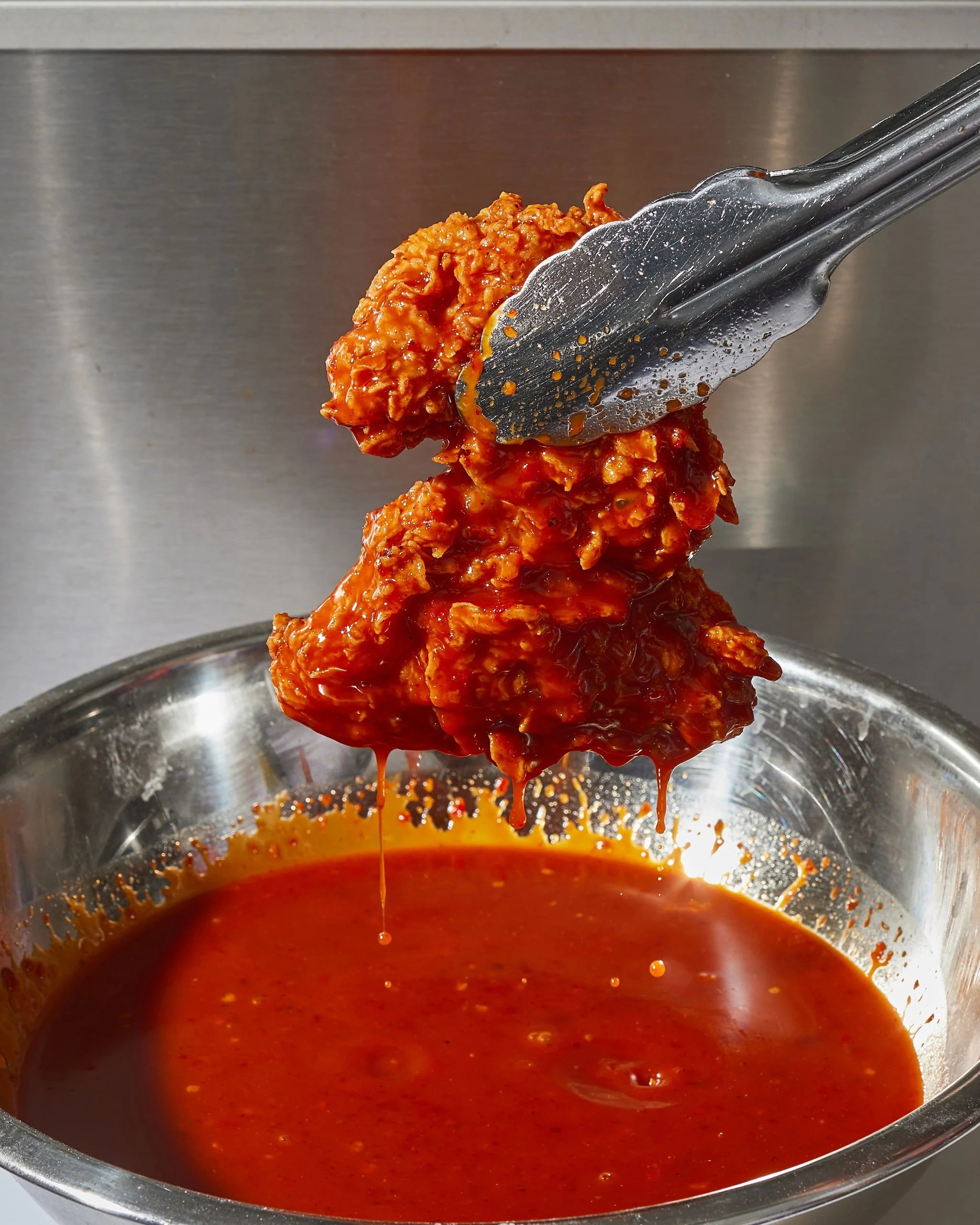 Tongs holding battered fried chicken coated in hot sauce above a metal bowl filled with hot sauce.