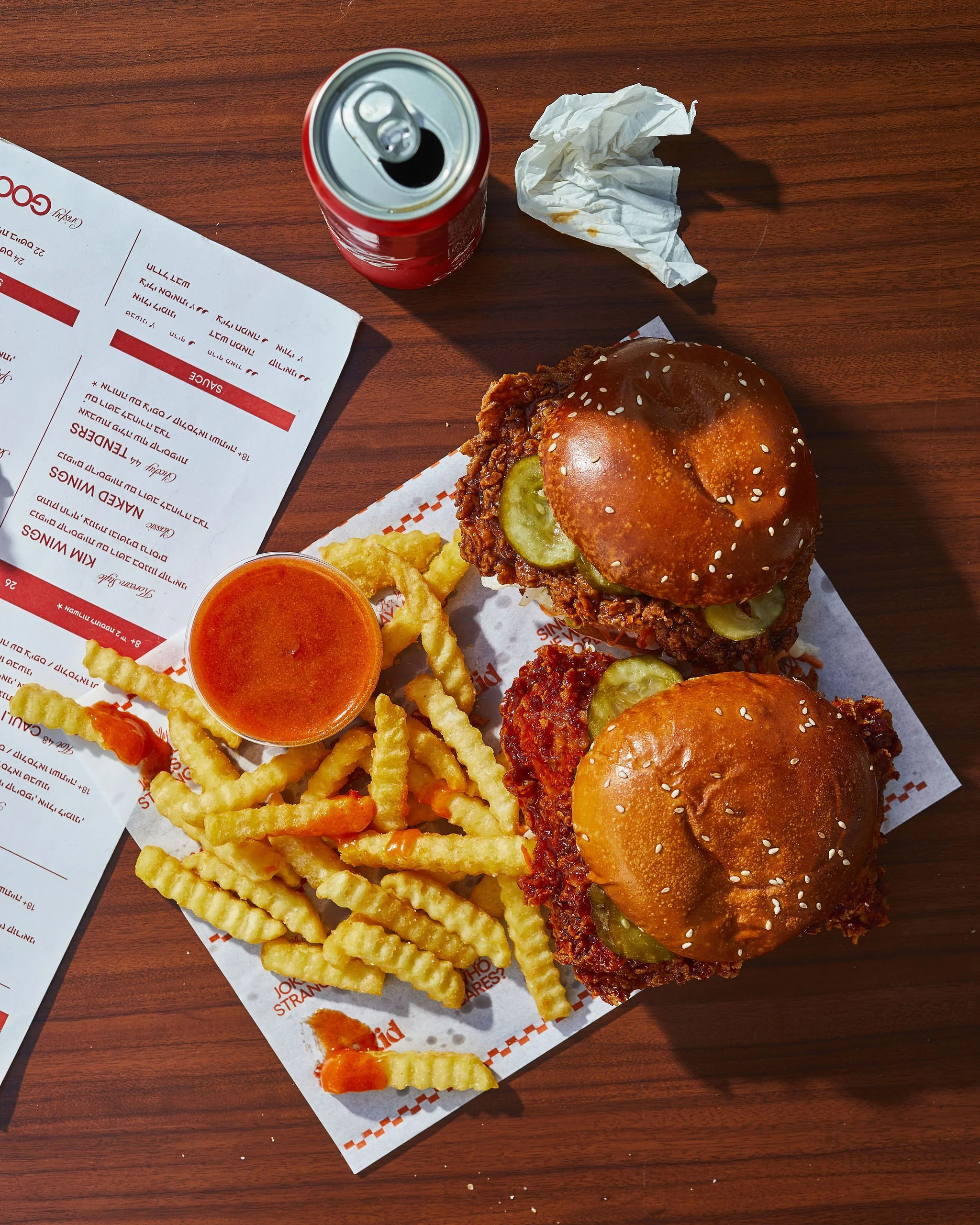 Two fried chicken sandwiches with pickles on sesame seed buns, crinkle-cut fries with a side of dipping sauce, a can of soda, a crumpled napkin, and a restaurant menu on a wooden table.