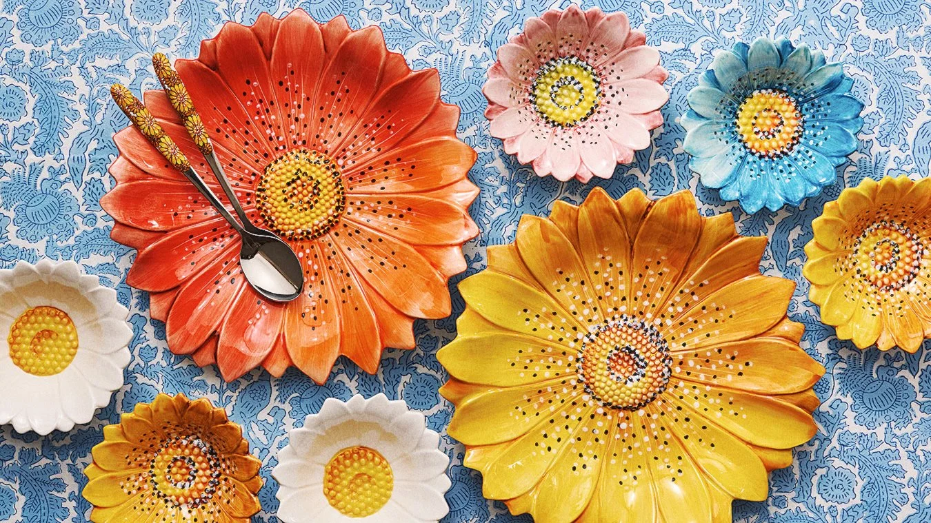 Colorful flower-shaped plates with dotted and beaded decoration arranged on a blue patterned tablecloth, some with spoons resting on them.