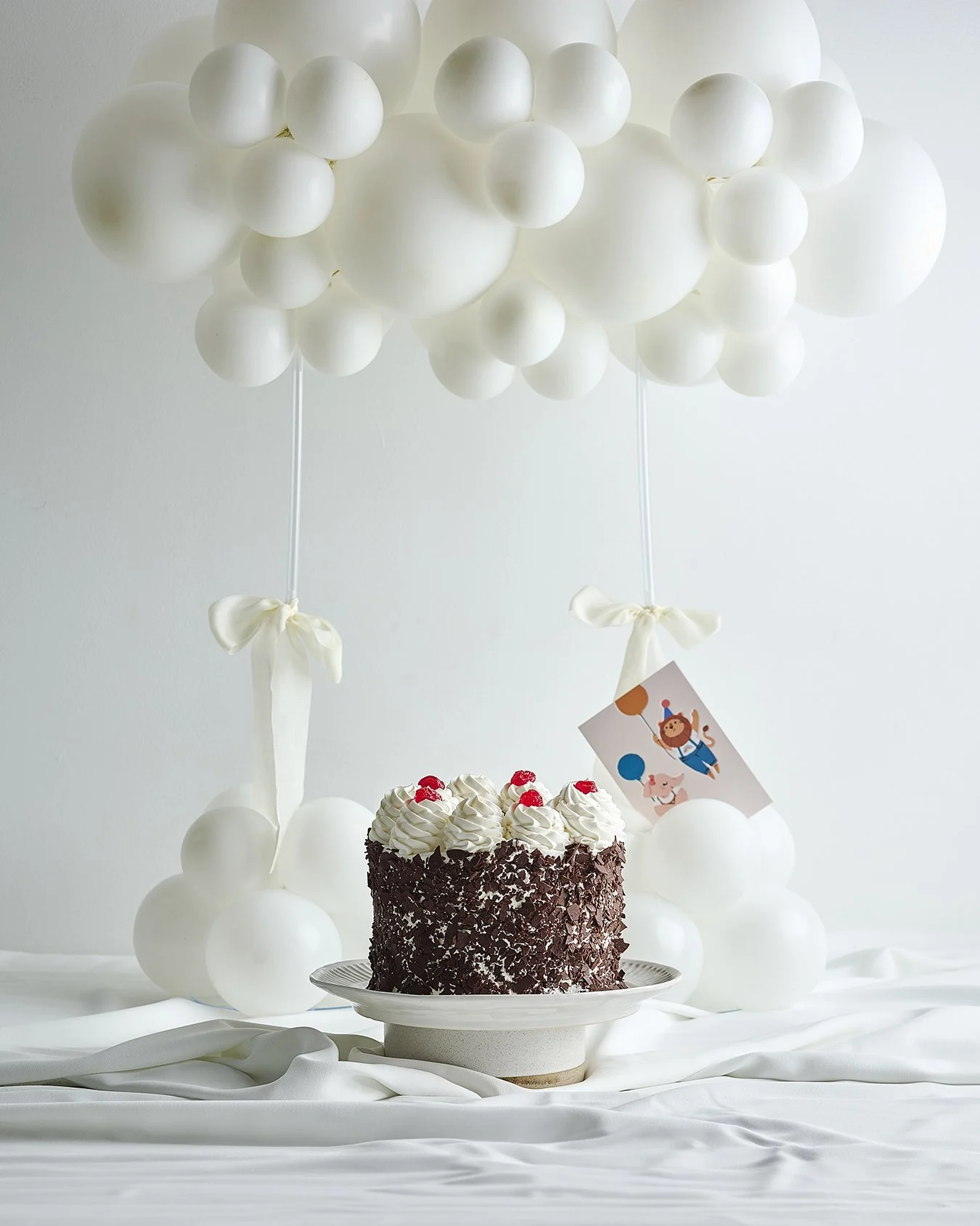 Chocolate birthday cake with whipped cream and cherries on top, surrounded by white balloons tied with ribbons, with a circus-themed invitation card attached to one balloon, all on a white draped surface.