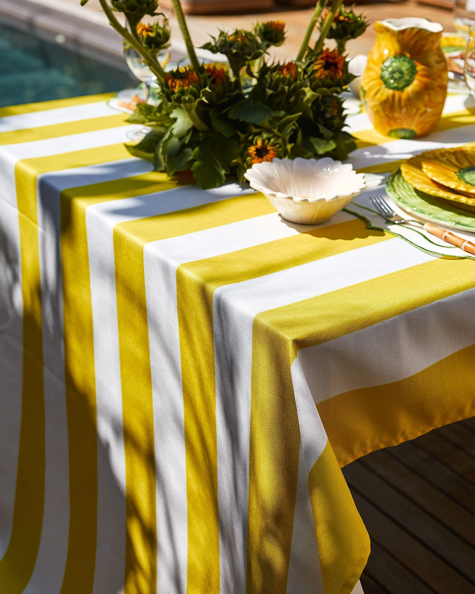 A table decorated for an outdoor event with a yellow and white striped tablecloth, a floral centerpiece of sunflowers, and various plates, cups, and utensils.