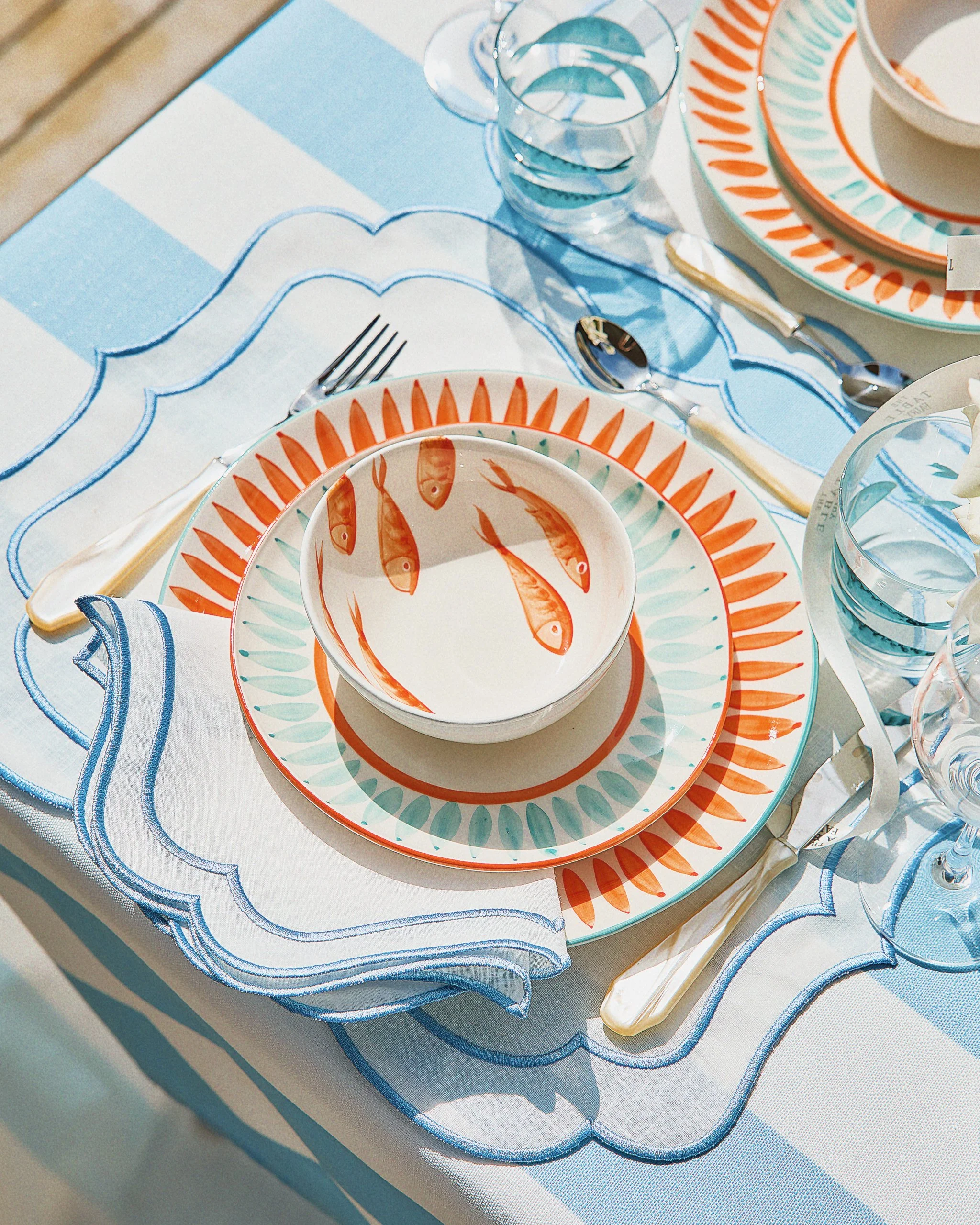 Table setting with fish-themed dinnerware, glassware, and cutlery on a light-colored table with blue and white decorative placemats.
