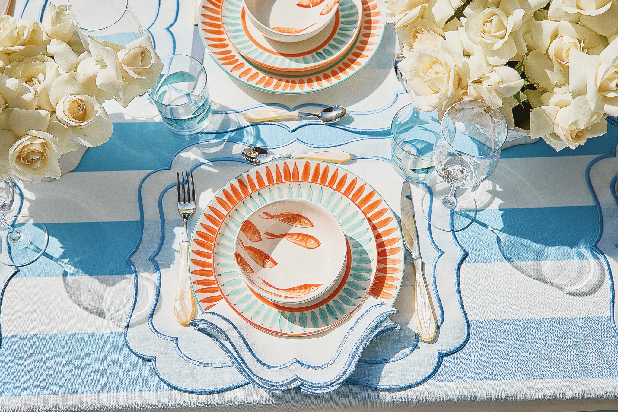 A table setting with fish-themed plates, surrounded by white roses and glassware, on a blue and white striped tablecloth.
