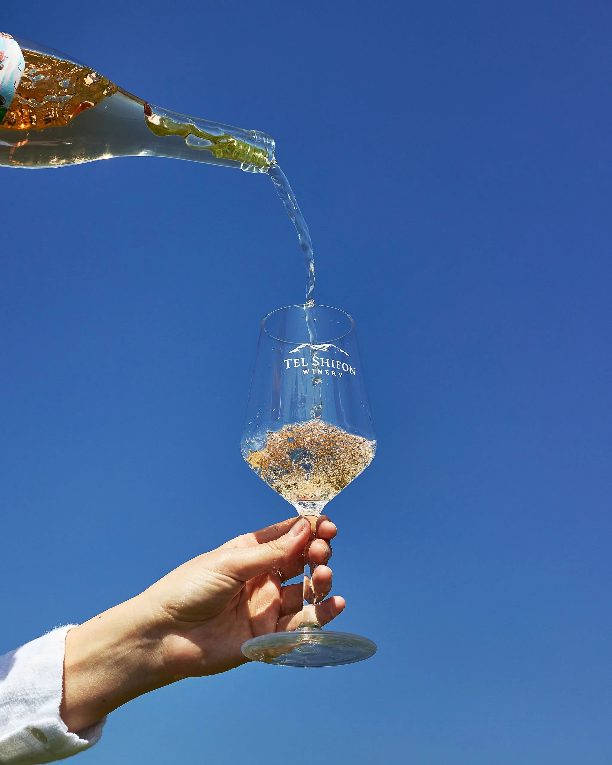 A hand holding a wine glass labeled 'TEL SHIFON WINERY' as white wine is being poured into it from a bottle against a clear blue sky.