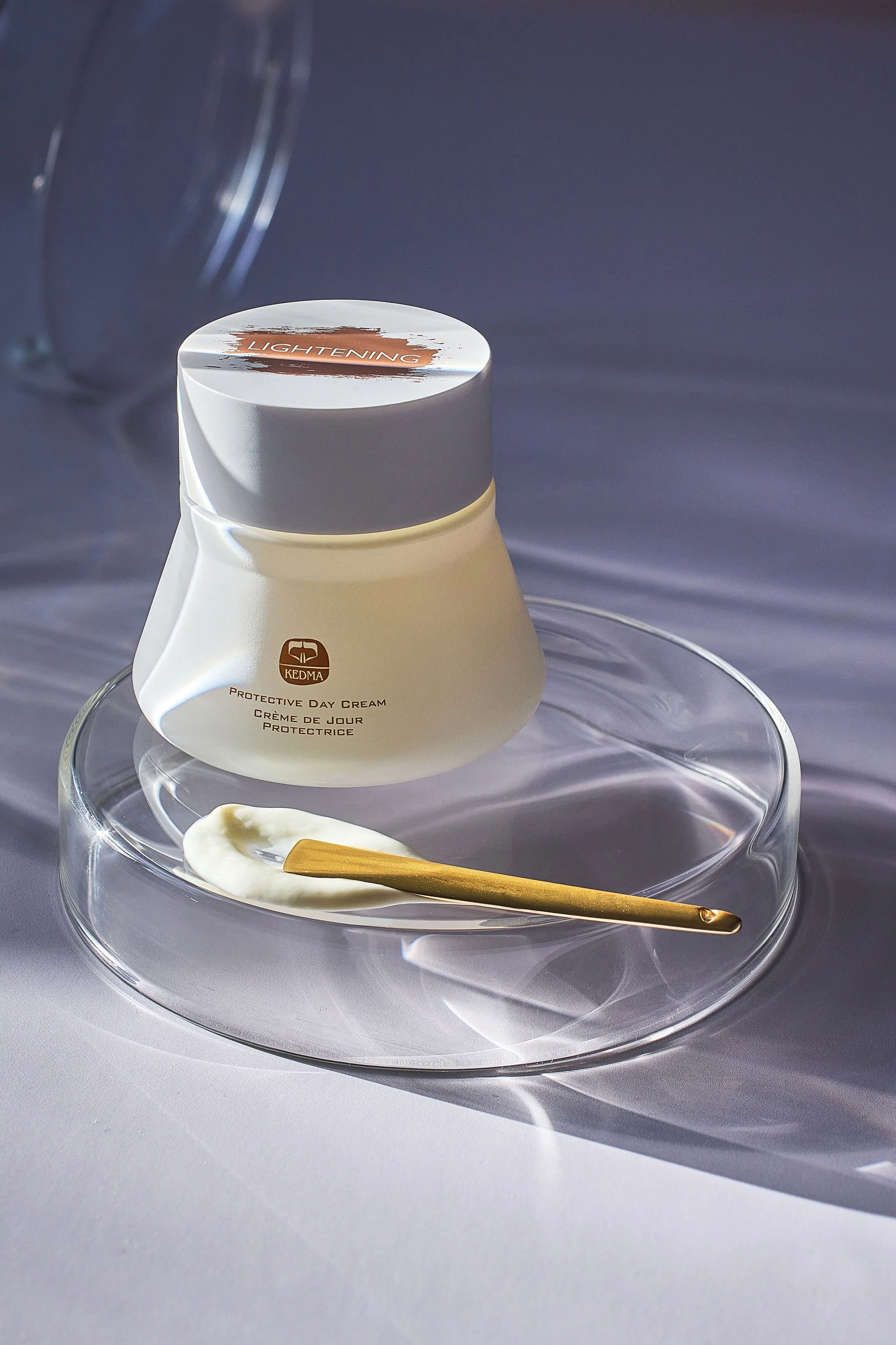 A white jar of Kedma protective day cream with a gold spatula and a smear of cream on a clear glass plate, set against a gray background.