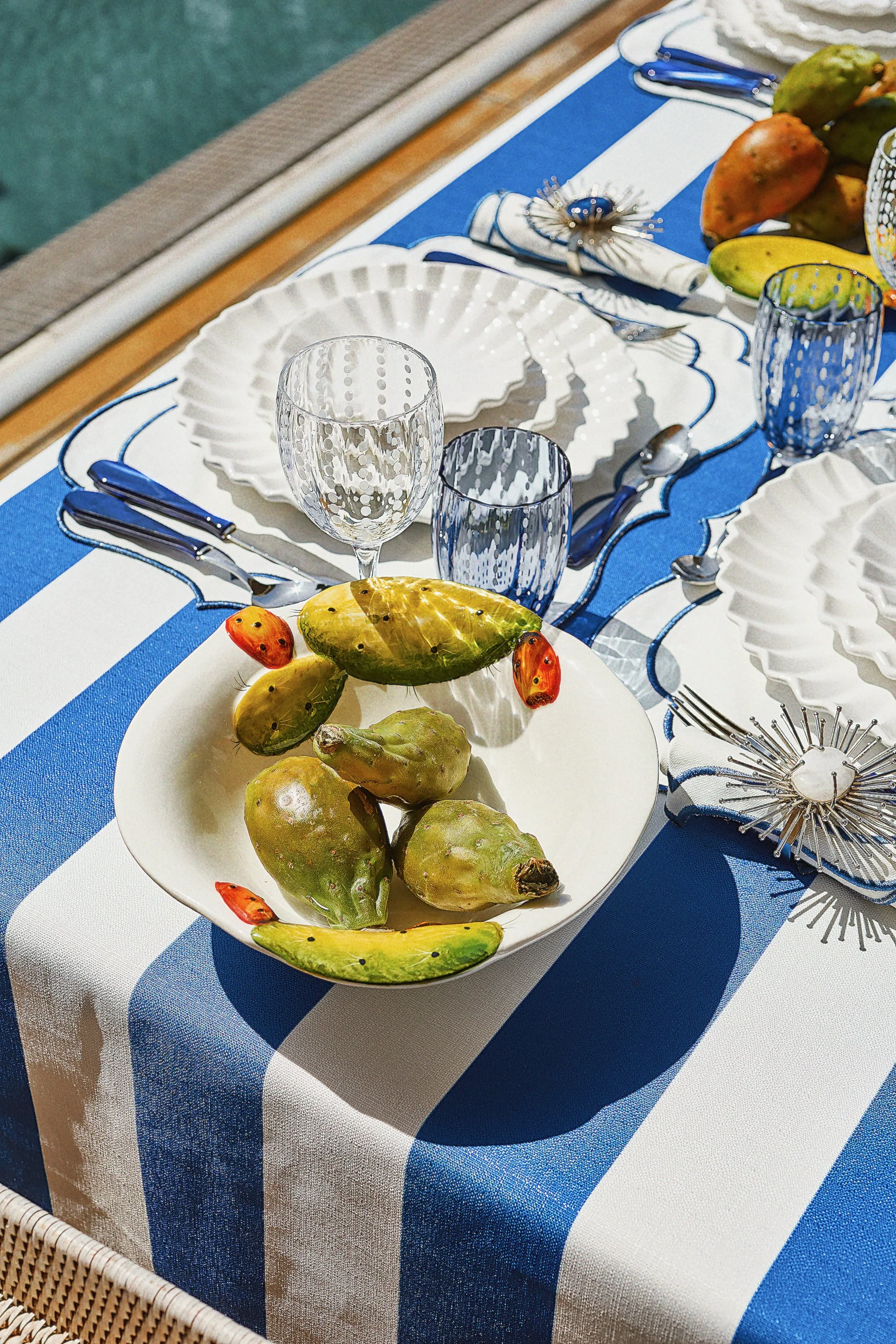 A table set with blue and white striped tablecloth, white and textured glassware, white plates with a scalloped edge, and a decorative silver candle holder. A white bowl holds prickly pears and decorative strawberries on a porch or outdoor dining are