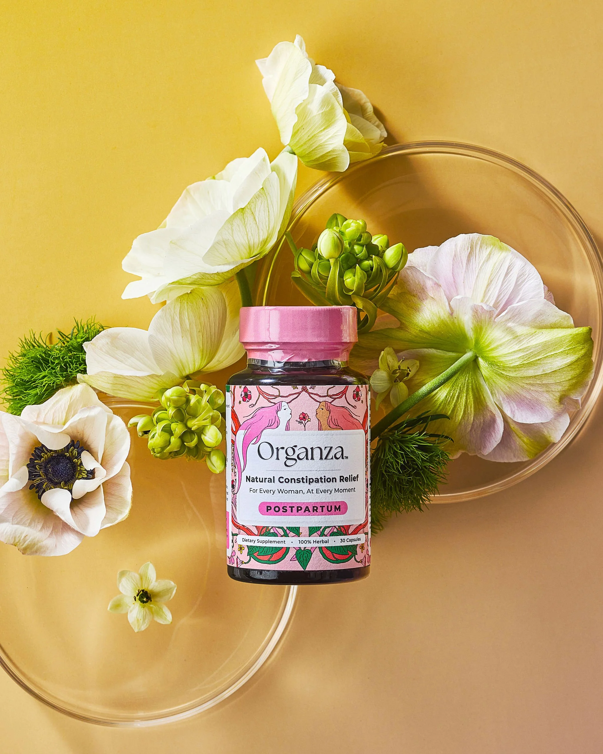 A bottle of Organza Postpartum Natural Constipation Relief supplement surrounded by white and green flowers and placed on a yellow background.
