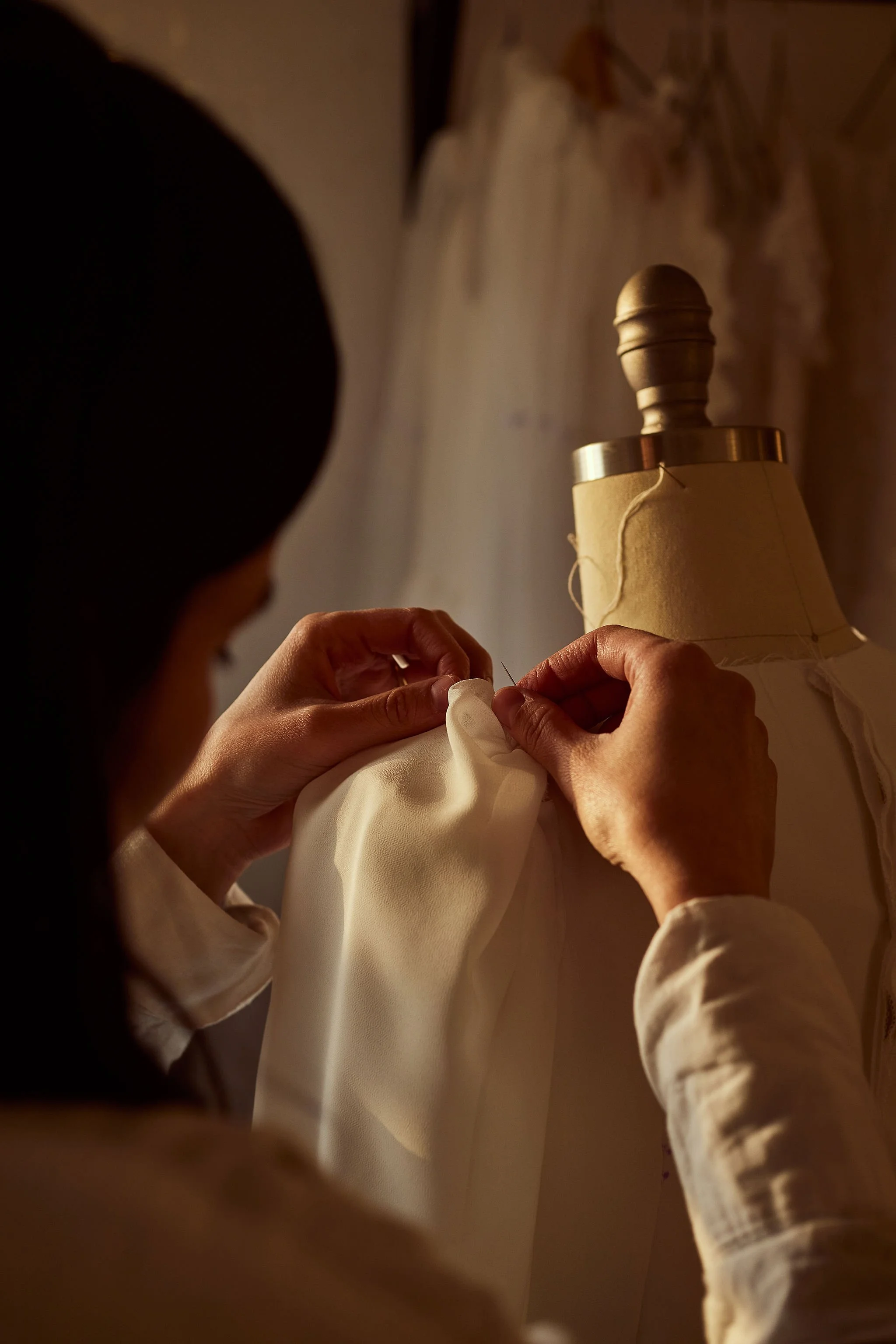 Person sewing a white fabric garment on a mannequin with a beige lampshade in the background.