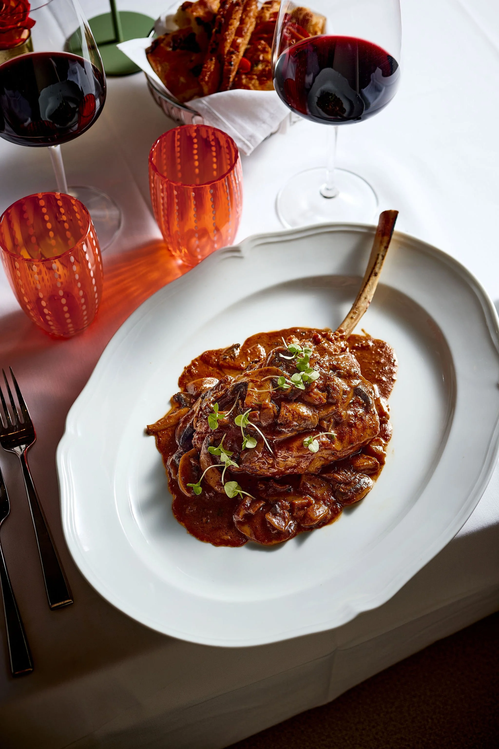 A plate of beef stew with a bone in a white decorative bowl, garnished with microgreens, on a dining table with two glasses of red wine and red patterned glasses, with a basket of bread in the background.