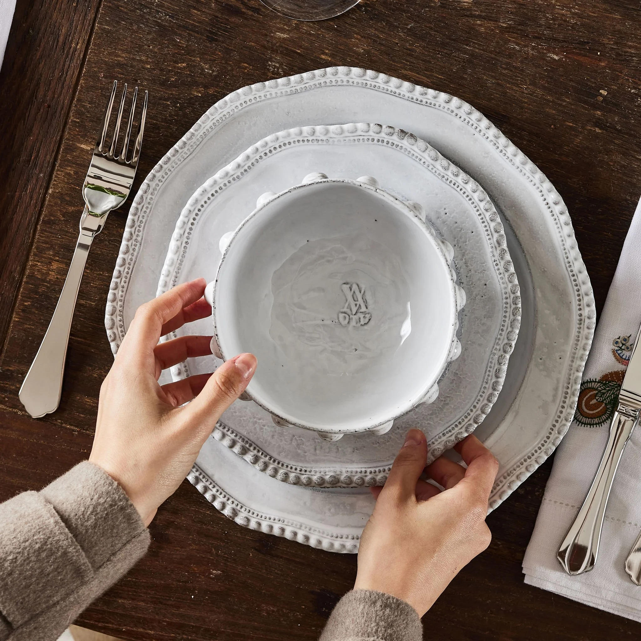 A table setting with white ceramic plates, a bowl, and utensils, with a person's hands arranging the tableware on a dark wooden table.