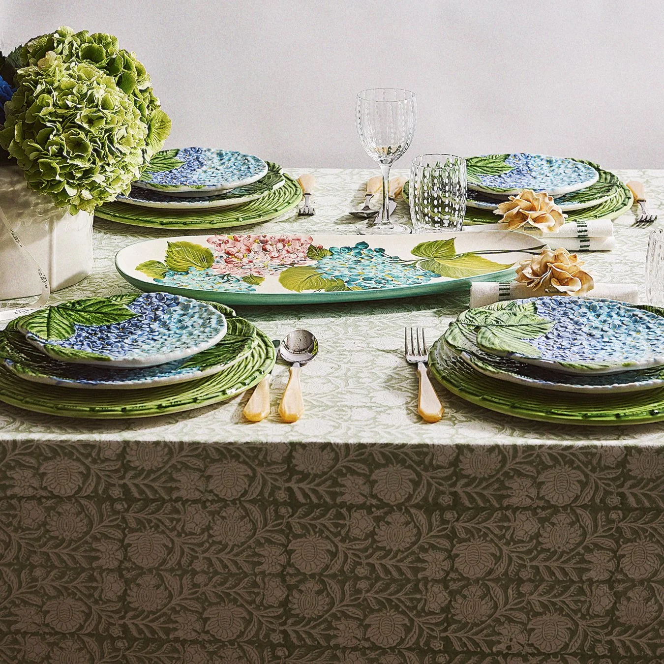 A table set with floral-patterned plates, silverware, wine glasses, and a large green flower arrangement.