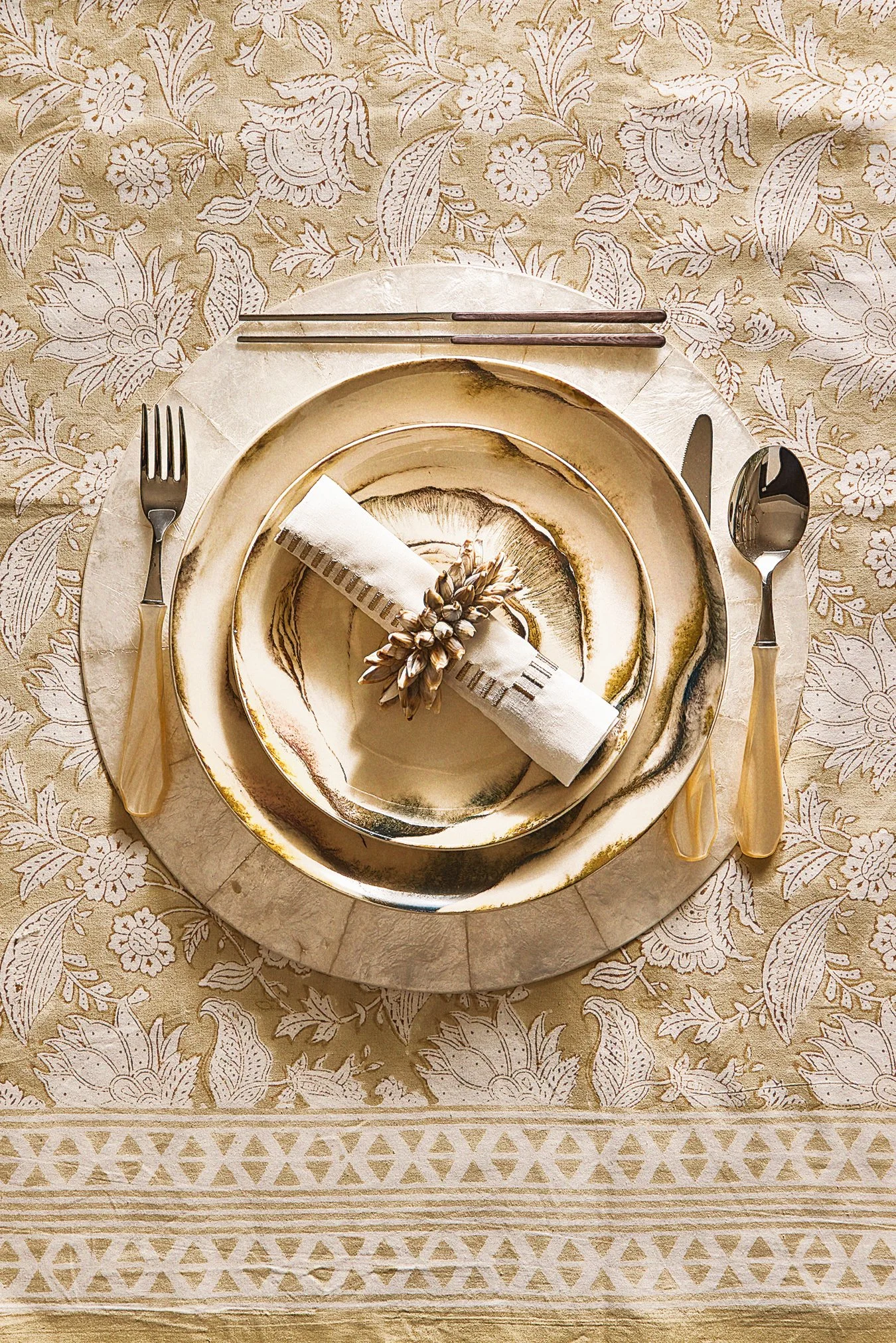 Table setting with a gold and cream marble plate, a smaller matching bowl, a rolled napkin with a decorative tassel and leaves, placed on a marble charger on a floral tablecloth with a fork on the left, a knife on the right, and chopsticks above.