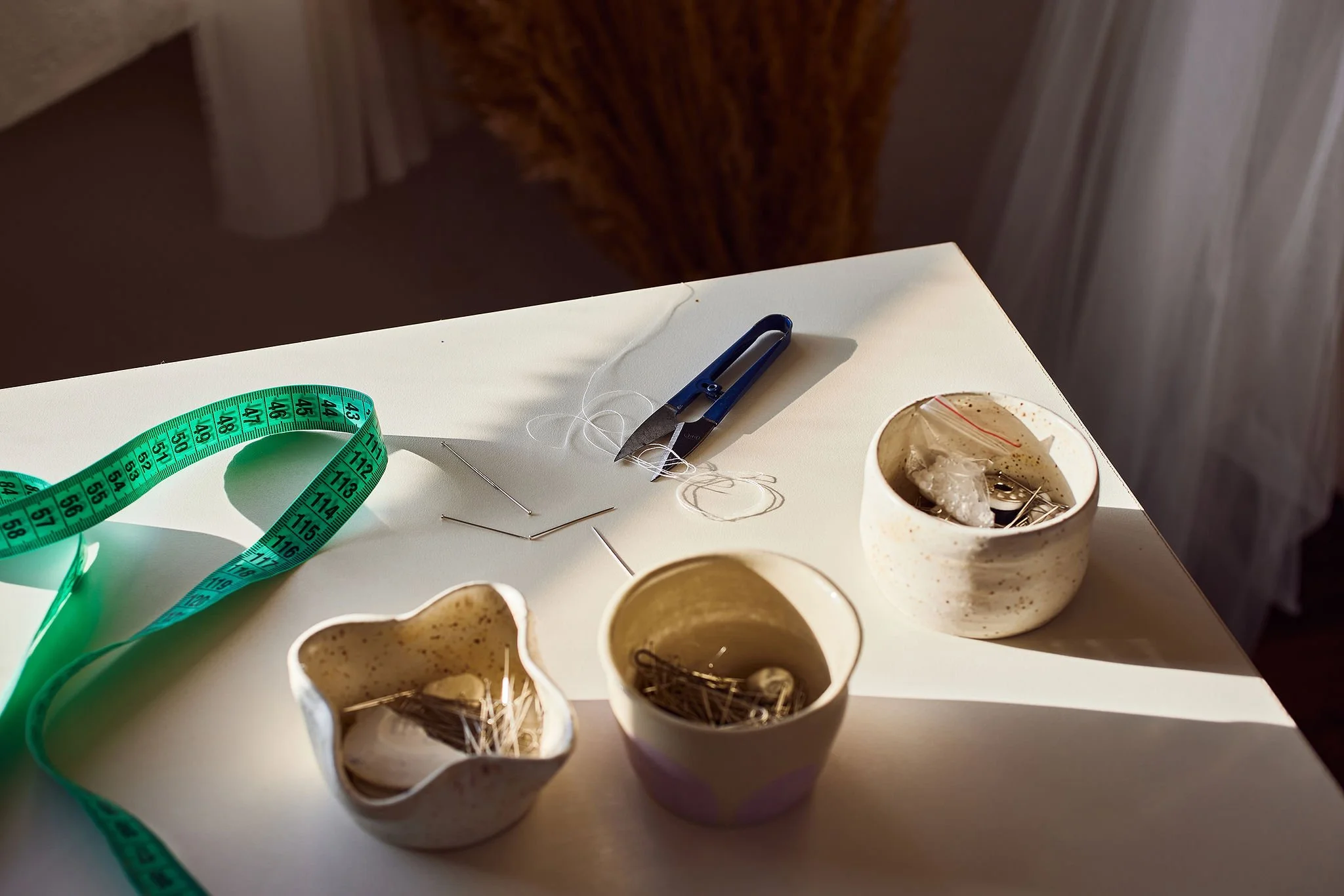 Sewing supplies on a white table, including a blue seam ripper, a measuring tape, sewing pins, and three small ceramic bowls filled with sewing notions.