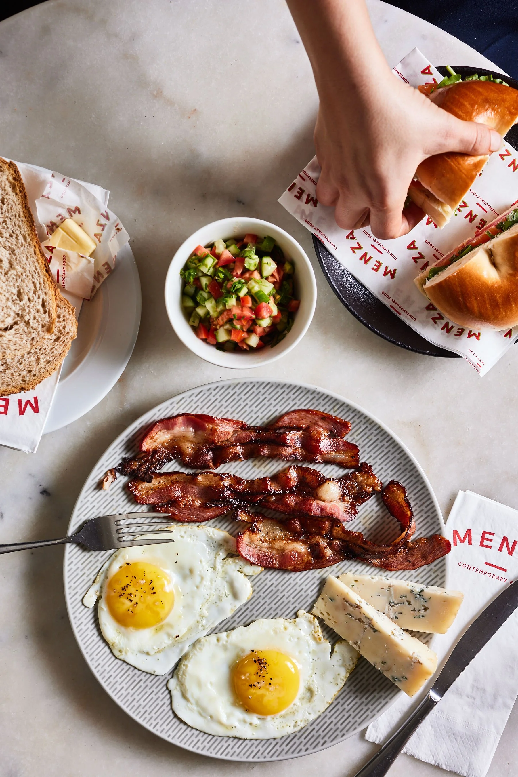 A breakfast plate with two fried eggs, crispy bacon strips, and blue cheese on a white plate. In the background, there's a bowl of chopped vegetables, a sandwich on whole wheat bread wrapped in MEZZANINE branded wrapping, and a hand holding a sandwic