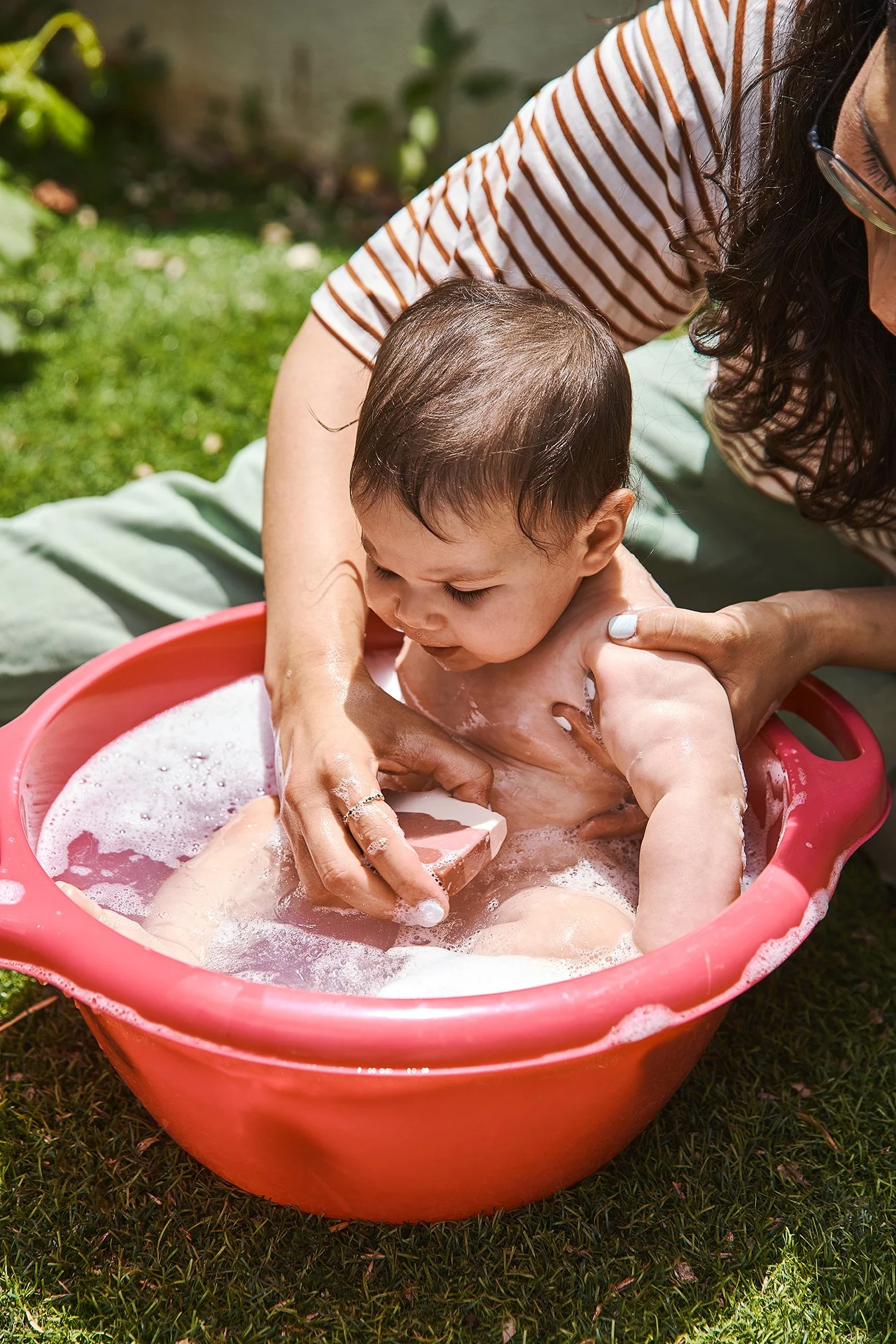 A woman giving a baby a bath in a pink plastic tub outside on grass, with bubbles and sunlight.