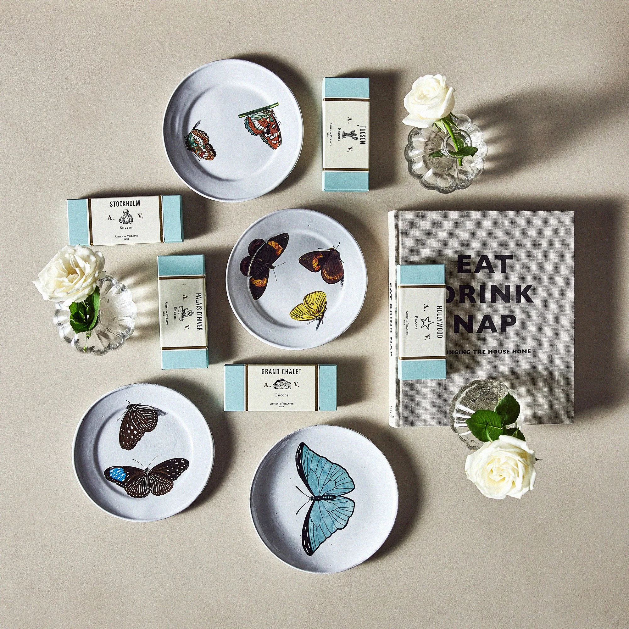 Table setup with butterfly and moth themed plates, small vases with white roses, and a book titled 'Eat Drink Nap' on a neutral background.