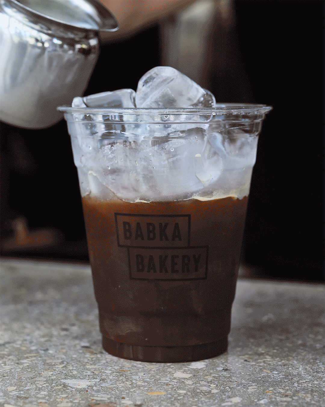 Plastic cup filled with iced coffee and ice cubes, with a Babka Bakery logo on the cup.