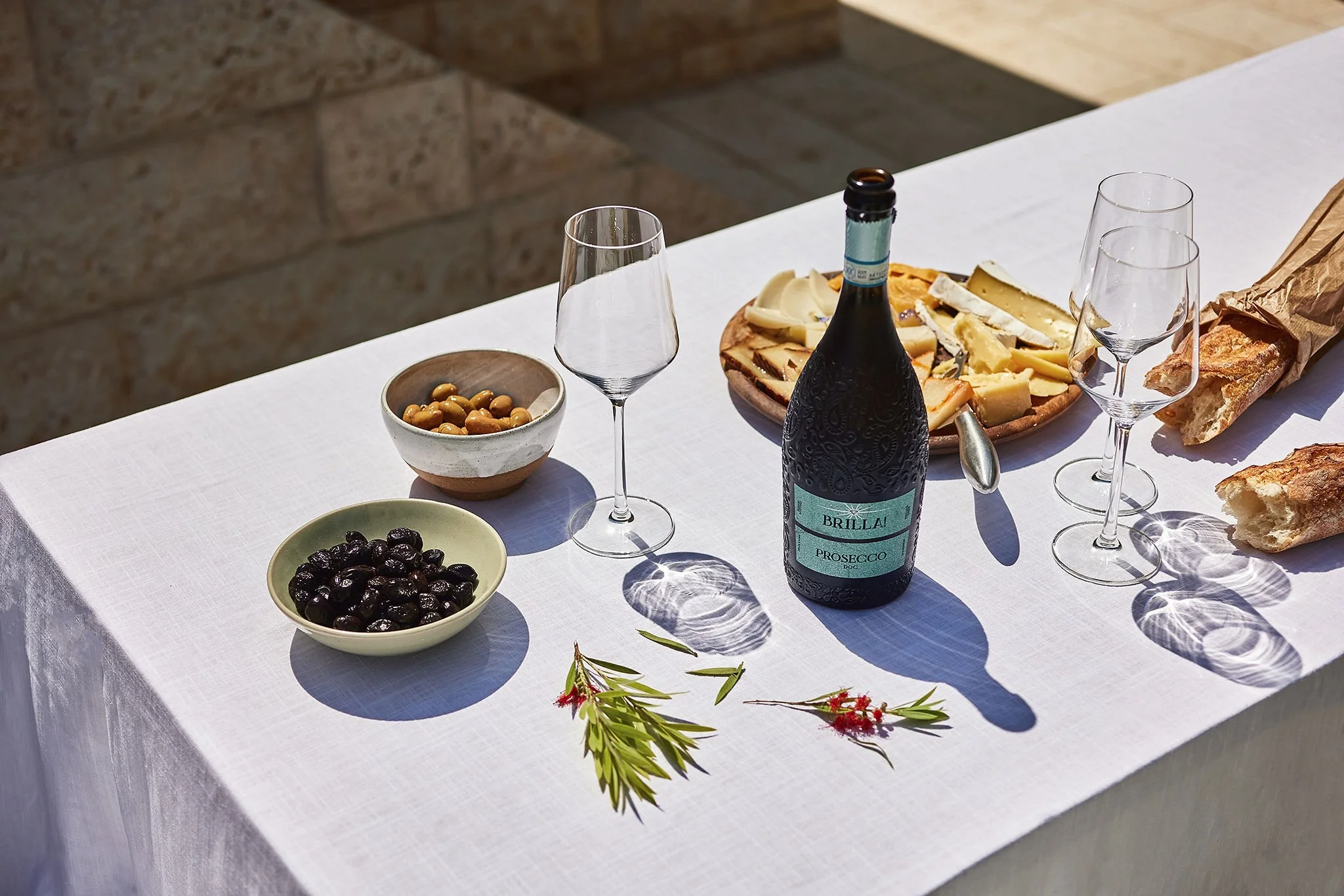 Table set with wine, glasses, cheese platter, bread, olives, and decorative sprigs on a white tablecloth.