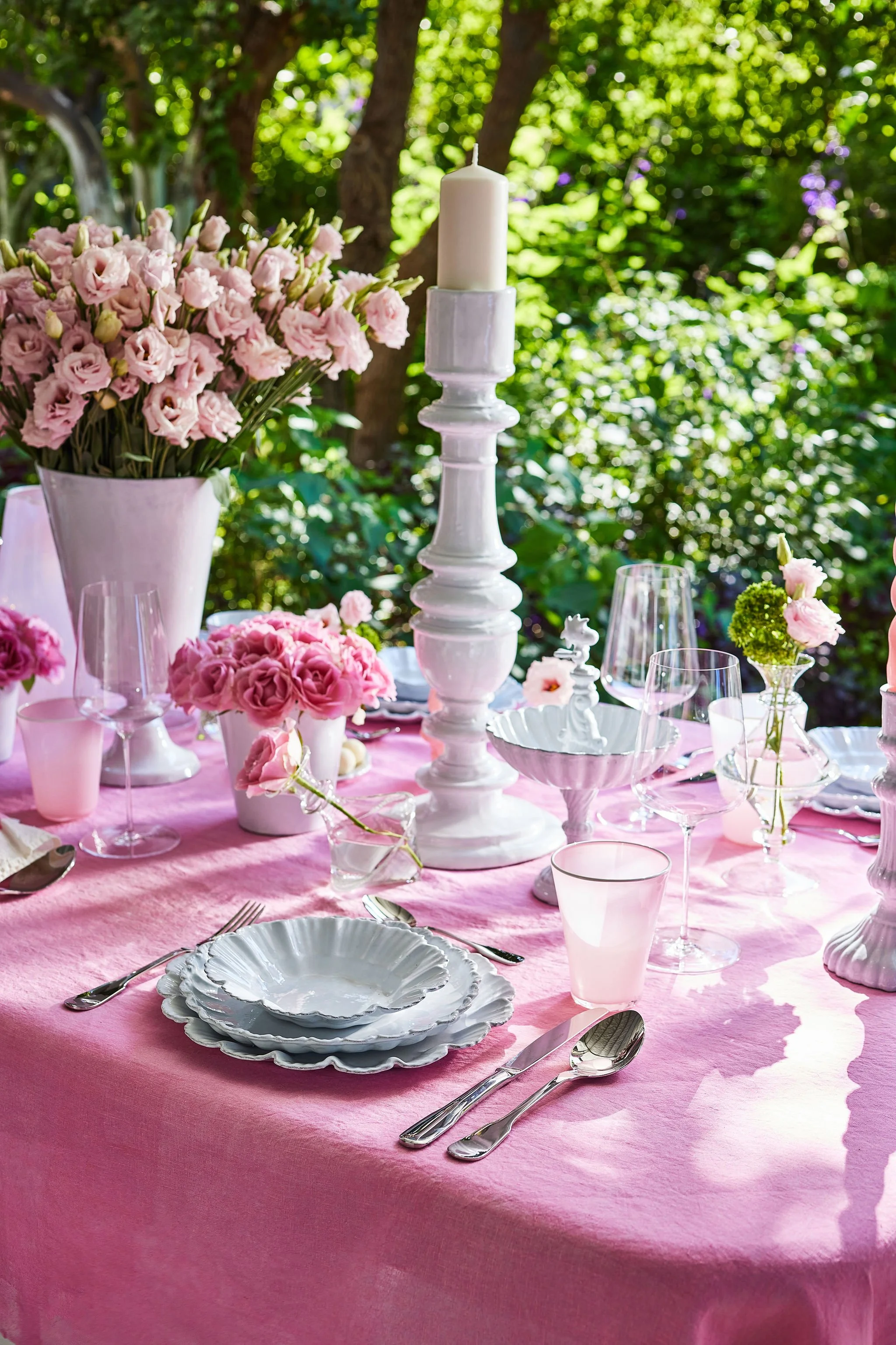 A pink tablecloth-covered table set for a meal, decorated with pink flowers in vases and a large white candlestick with a white candle, with trees and greenery in the background.