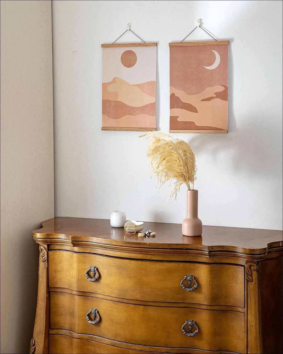 A wooden dresser with decorative handles, a pink vase with pampas grass, a small white cup, and some small decorative items on top, with pink and beige abstract landscape prints hanging above.