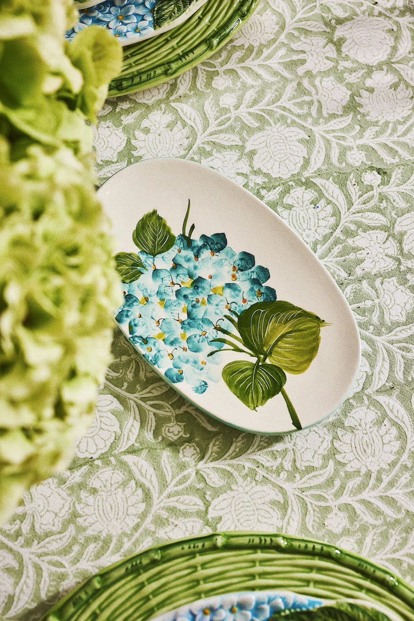Decorative oval plate with a floral design of blue hydrangeas and large green leaves, placed on a table with green patterned tablecloth. Part of a green wicker basket is visible at the bottom and a basket with blue and white flowers is at the top.