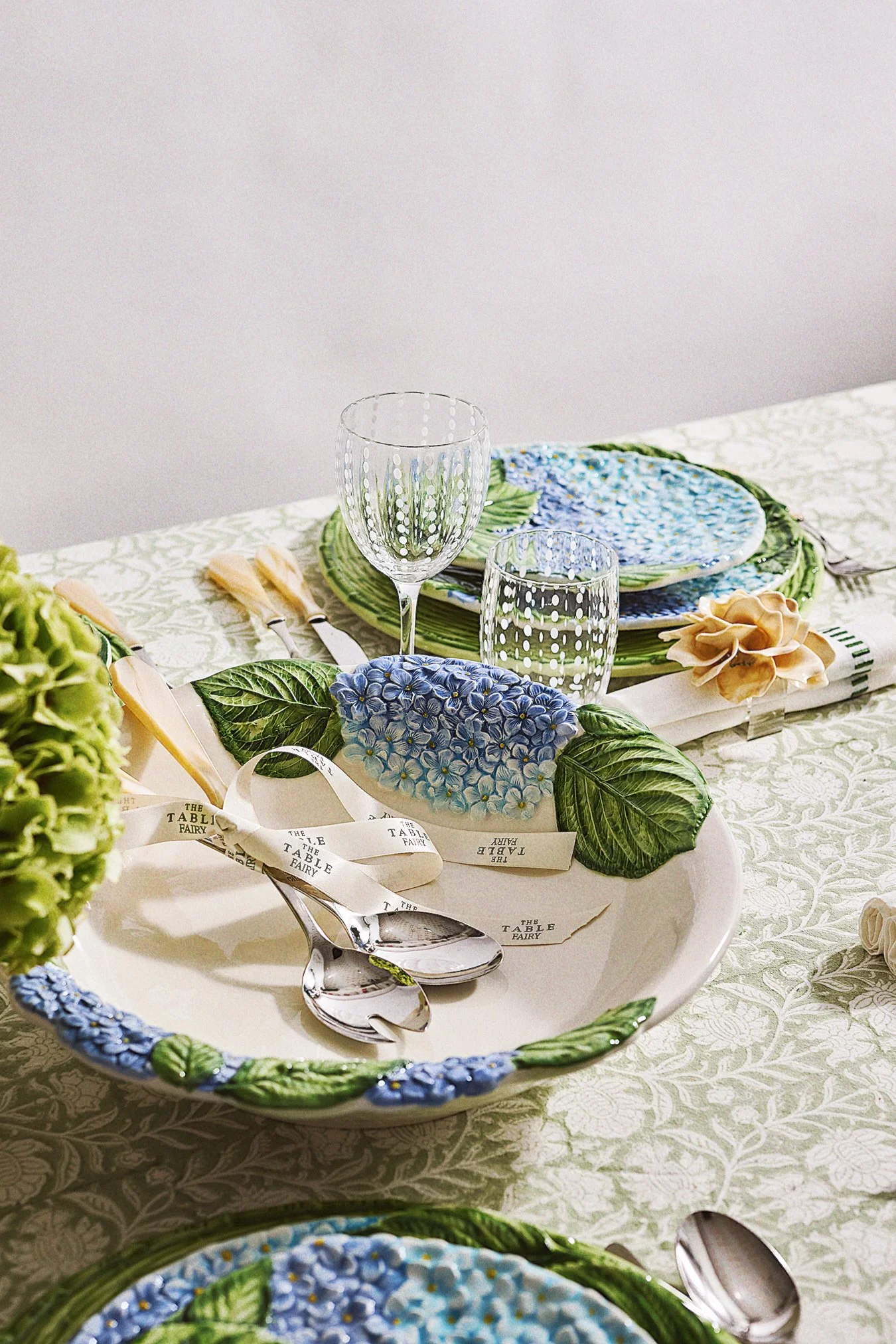 A table setting with floral patterned plates, green leaf-shaped dishes, and glassware. The tablecloth has a light floral design, and silverware is arranged on the table.