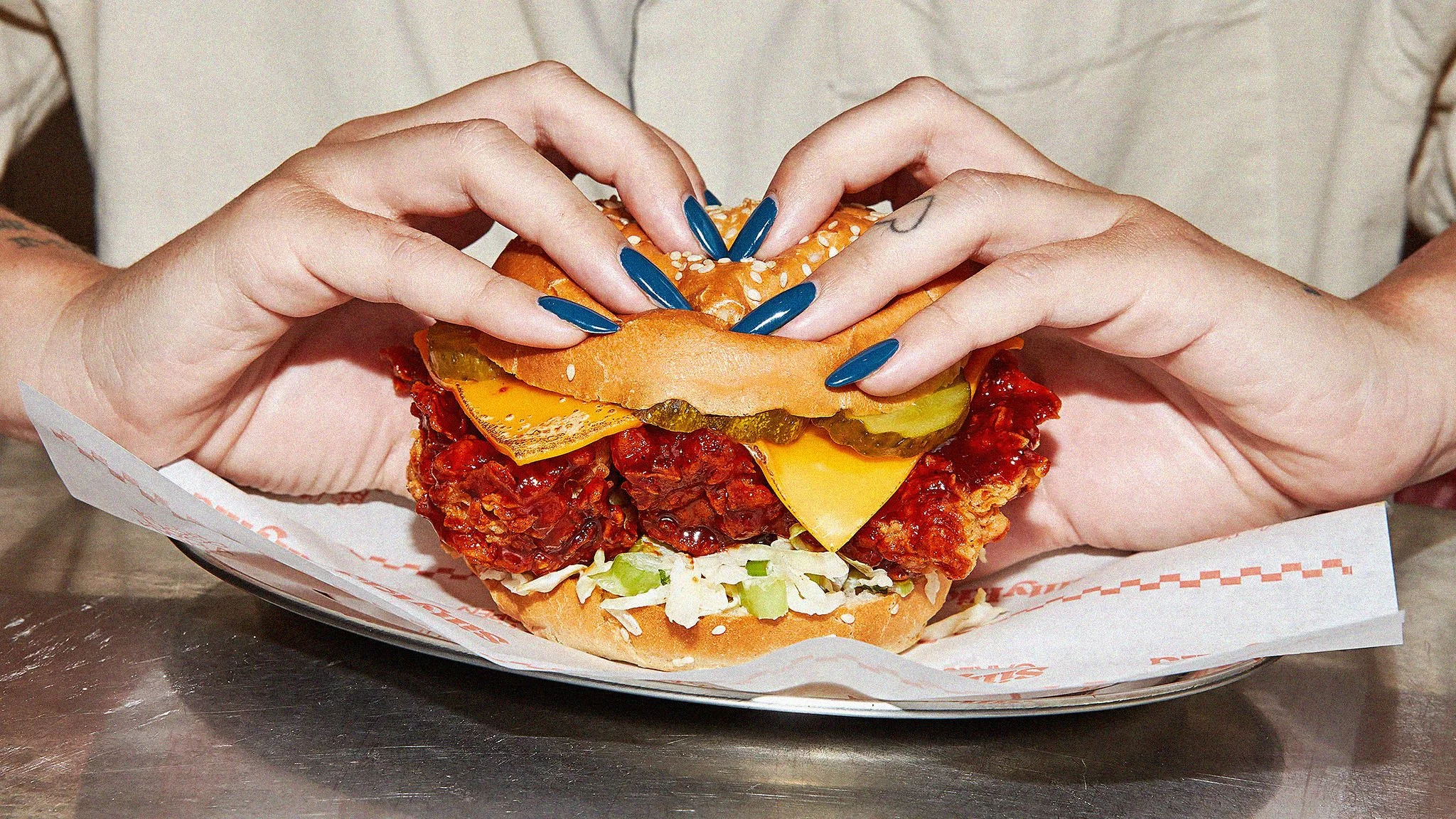 Person holding a large sandwich with pickles, cheese, shredded lettuce, chili, and a sesame seed bun.