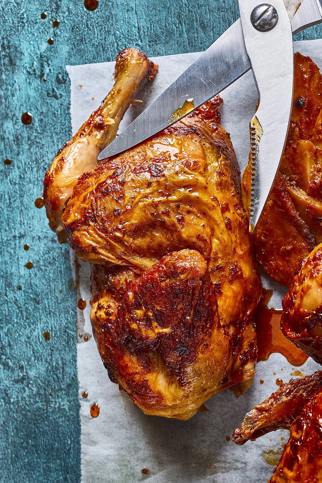 Close-up of a cooked chicken thigh with crispy skin, on parchment paper, with a meat fork piercing it, on a distressed blue wooden surface
