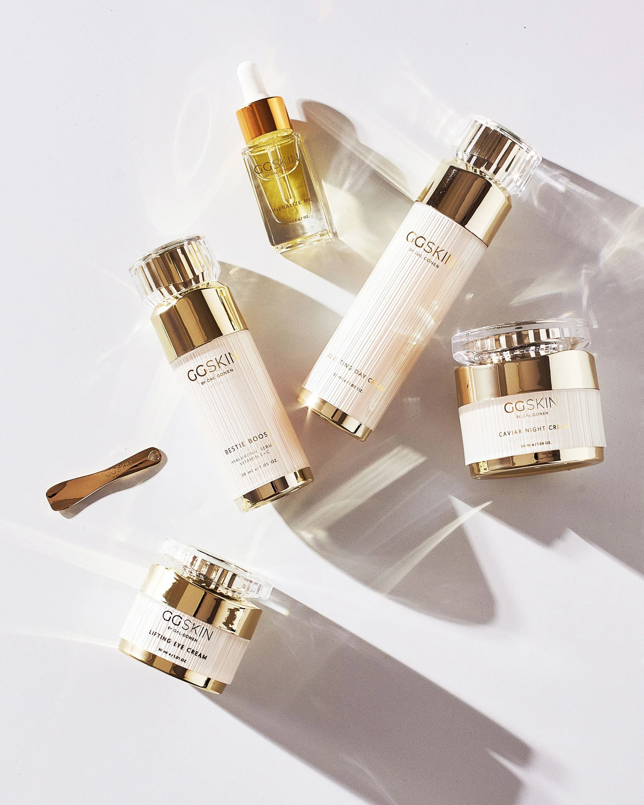 A collection of skincare products on a white surface with shadows cast by sunlight. The products are primarily white and gold, including bottles of lotion, serum, eye cream, and a small jar of caviar night cream.