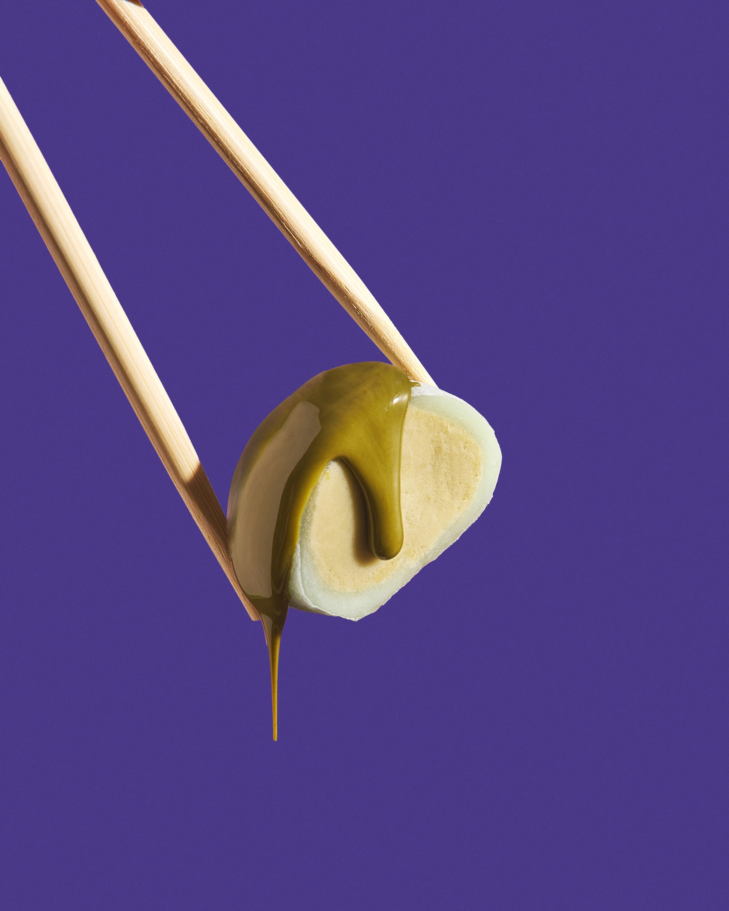 Close-up of a heart-shaped marshmallow with green caramel sauce drizzling down, held by wooden sticks against a purple background.
