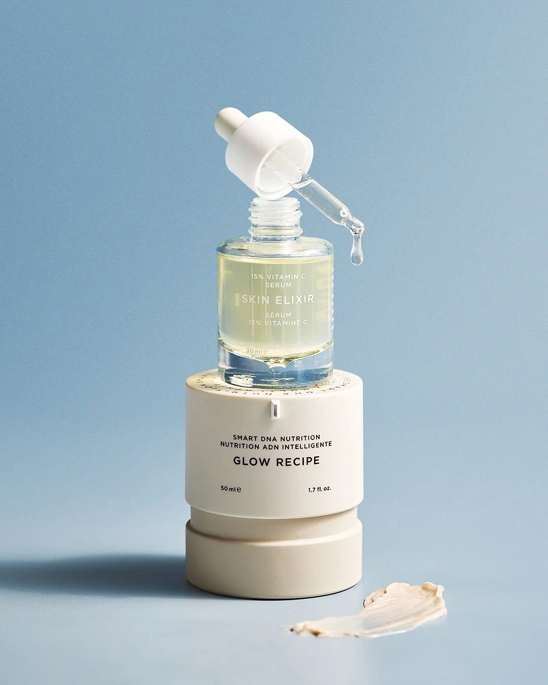 A skin care serum bottle with a dropper resting on top, a white container labeled 'Glow Recipe,' and a spatula with cream on it against a light blue background.