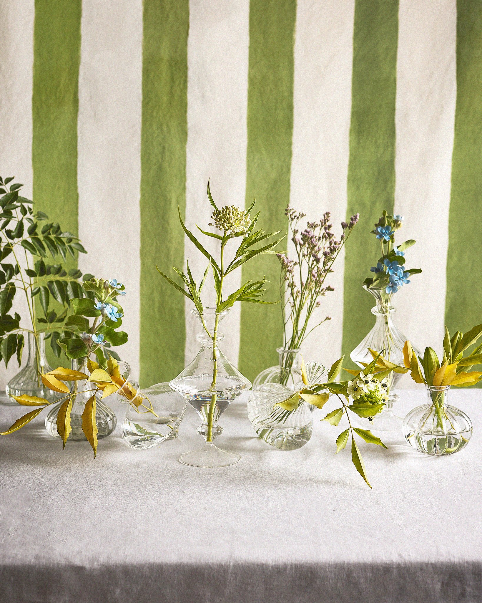Several small clear glass vases with green, purple, and blue flowers and greenery arranged on a white table, with a background of vertical green and white striped walls.
