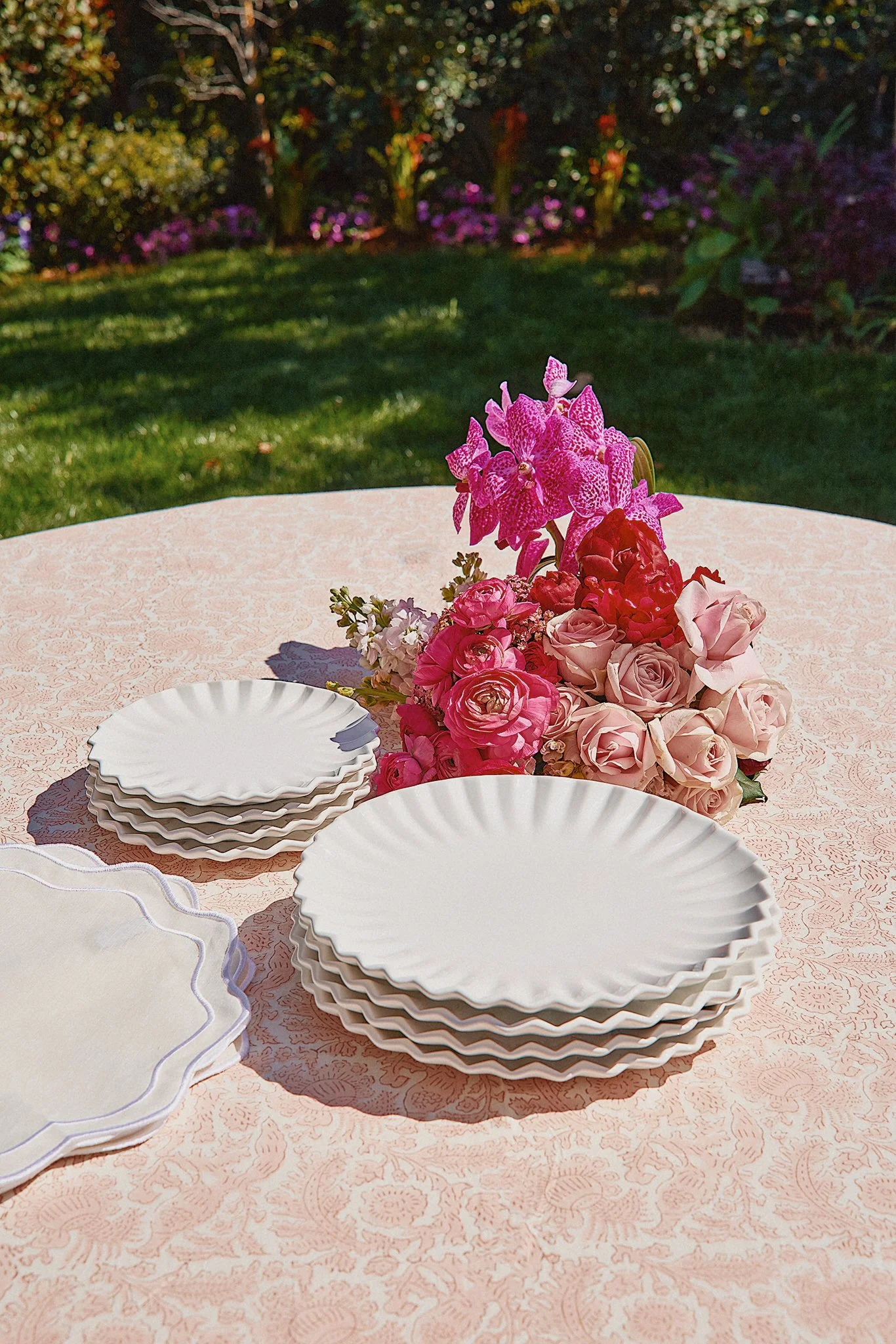 A round pink tablecloth-covered table set with white scalloped-edge plates and a floral centerpiece of pink and white roses with purple orchids outside in a garden.