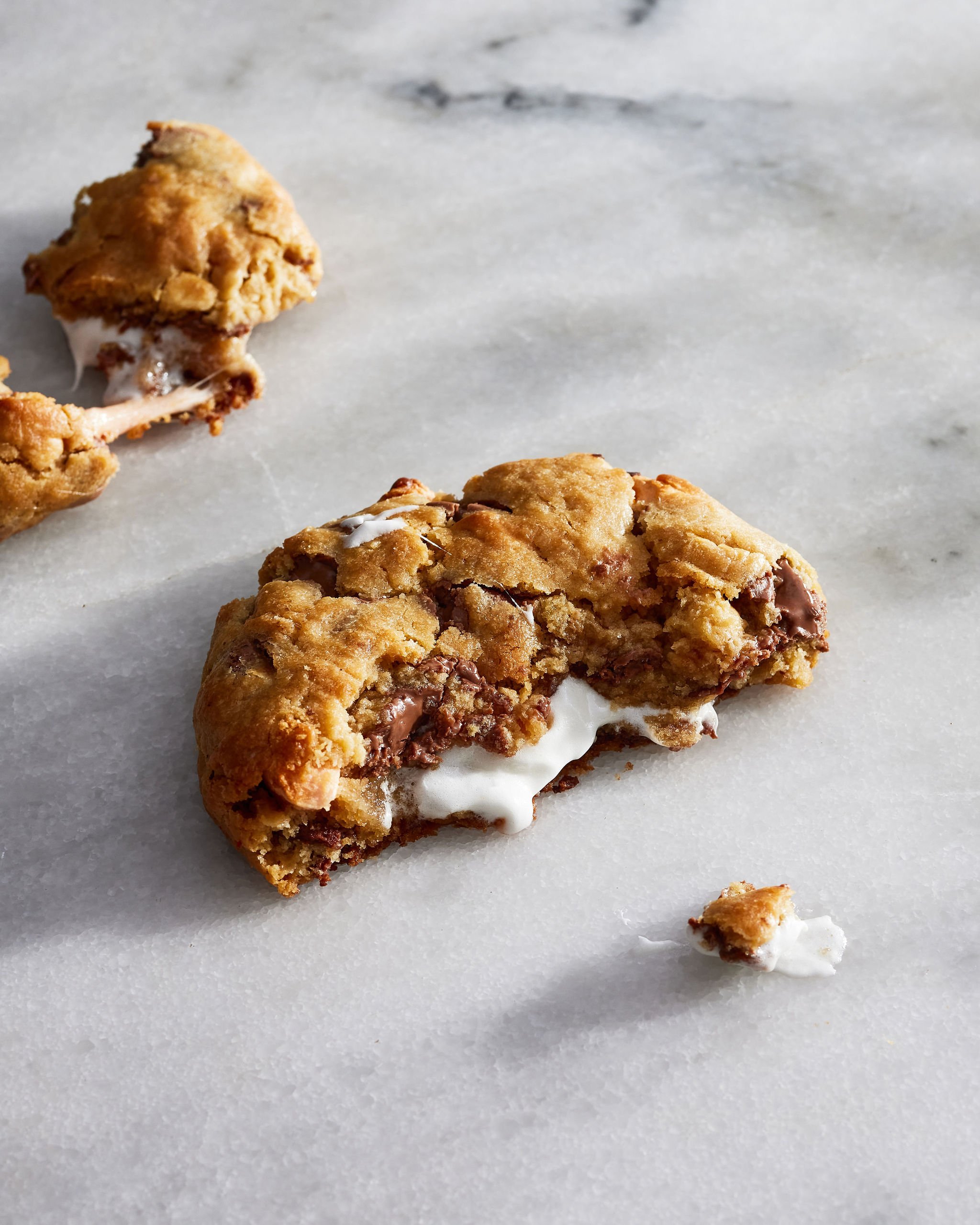 Close-up of a broken chocolate chip cookie with melting chocolate and white cream inside, on a white marble surface.