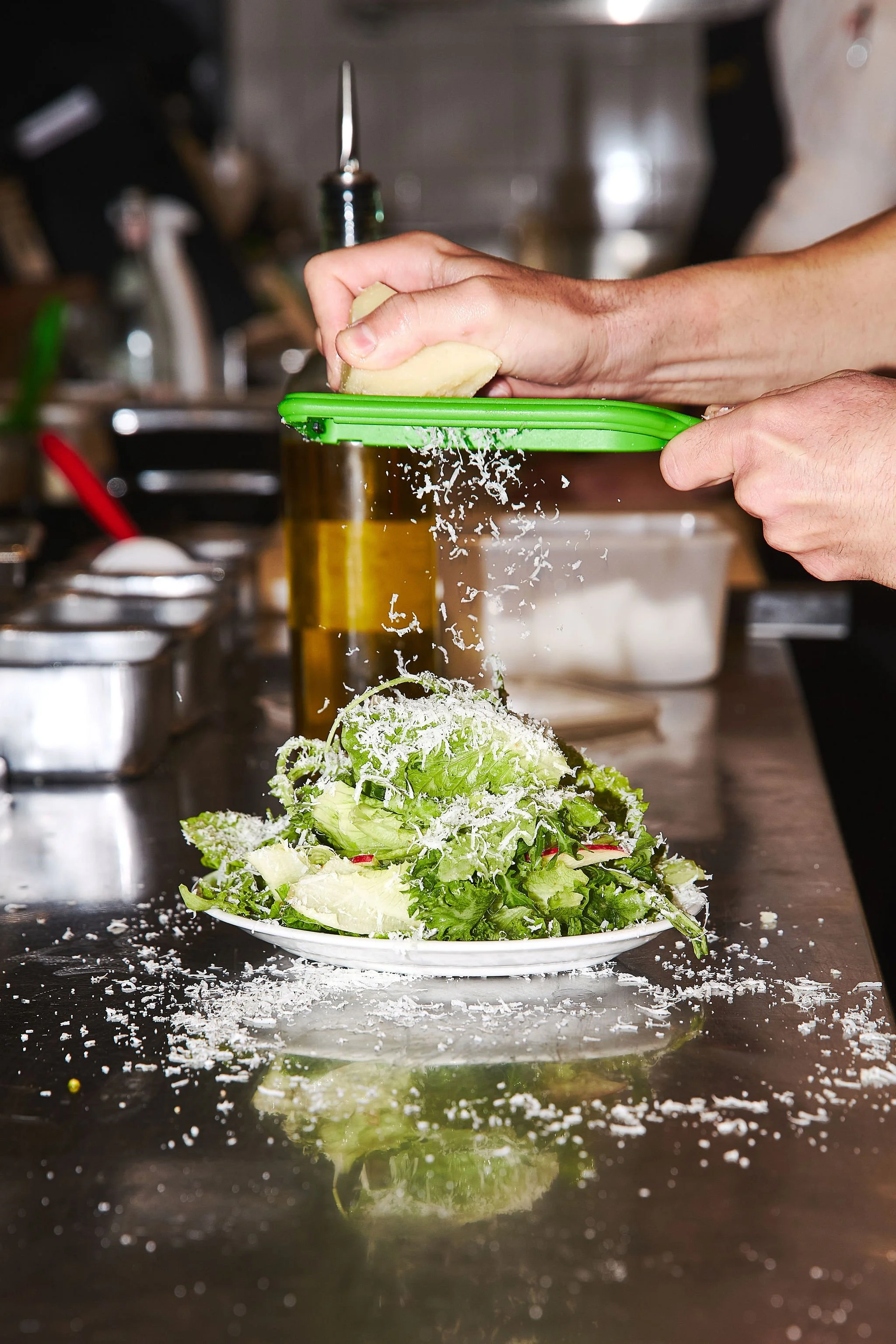Person grating cheese over a fresh salad on a white plate in a kitchen.