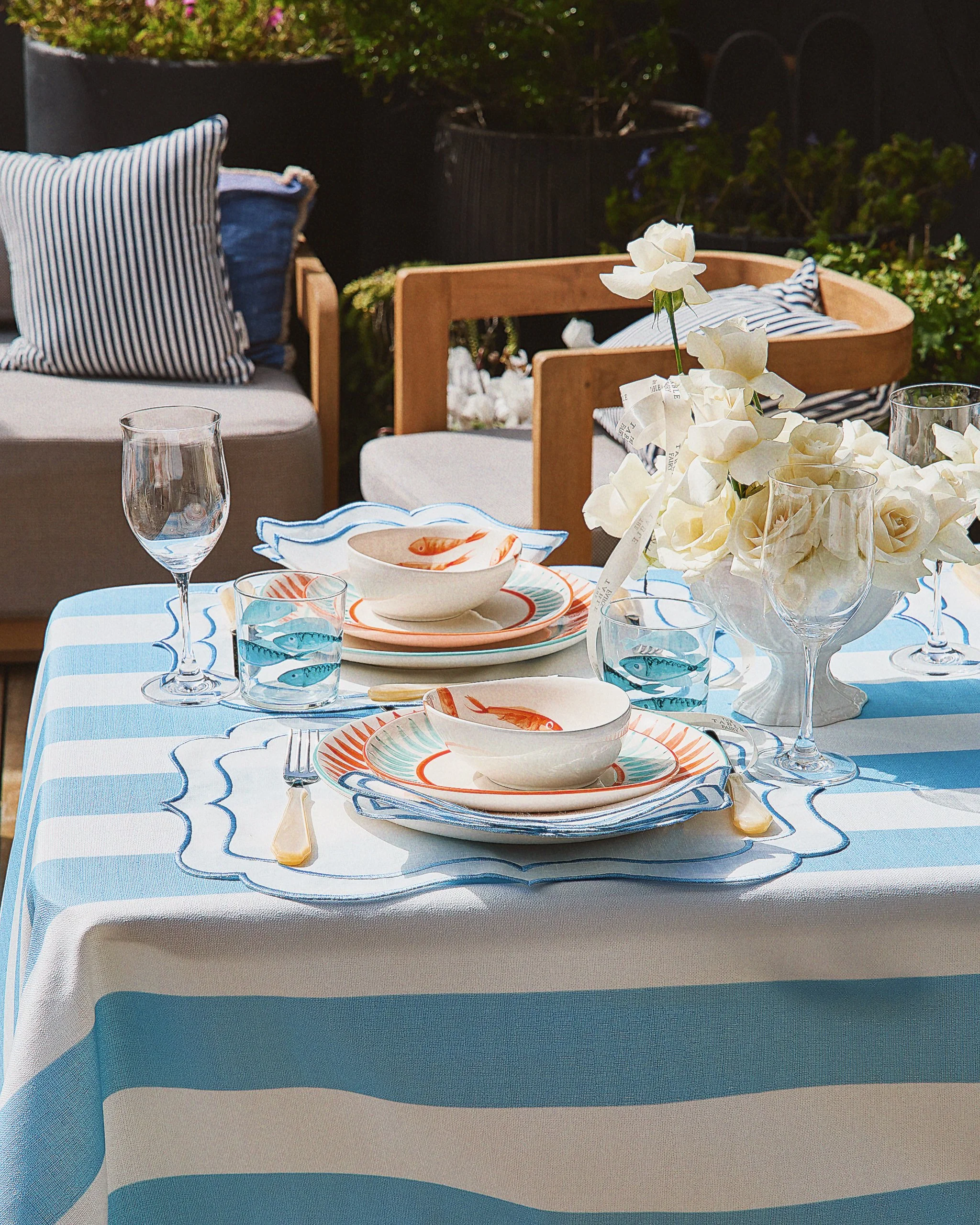Outdoor dining table with blue and white striped tablecloth, seafood-themed dishware, wine glasses, a floral centerpiece with white roses, and place settings with cutlery.