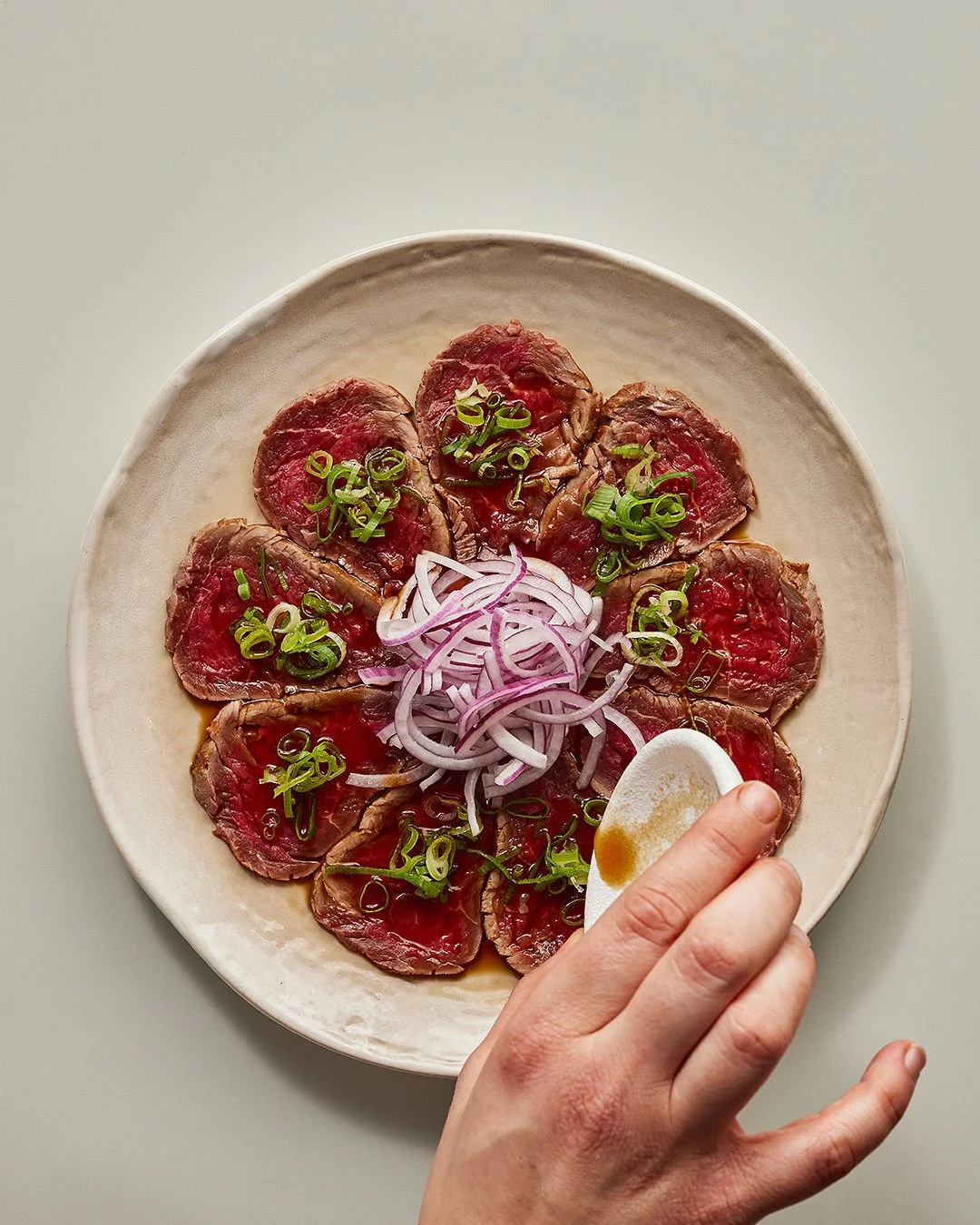 A plate of sliced raw beef carpaccio garnished with chopped green onions and thinly sliced red onions in the center, with a hand holding a small bowl of soy sauce or dressing nearby.