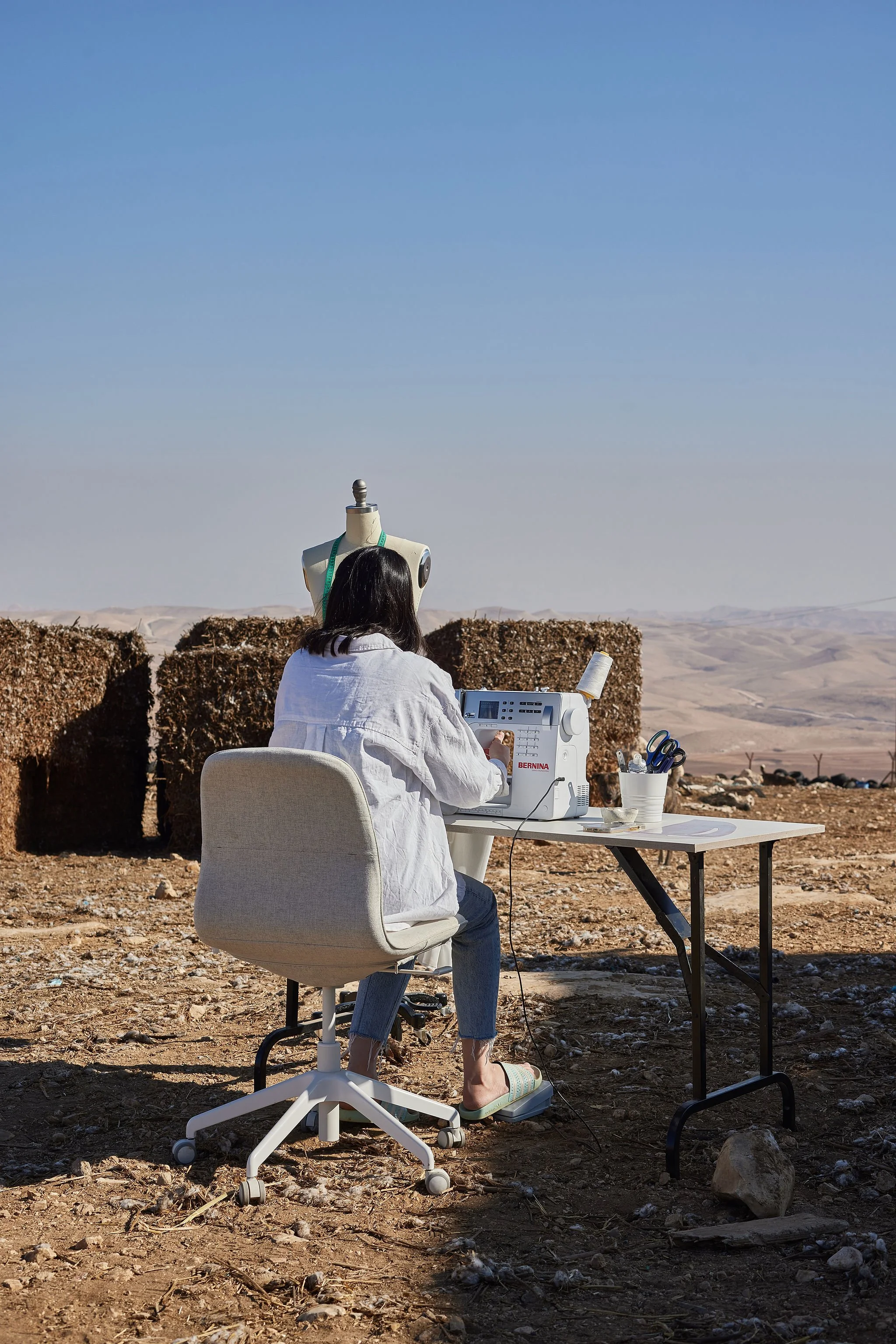 A woman sewing on a machine outdoors in a desert landscape, with a mannequin and desert hills in the background.
