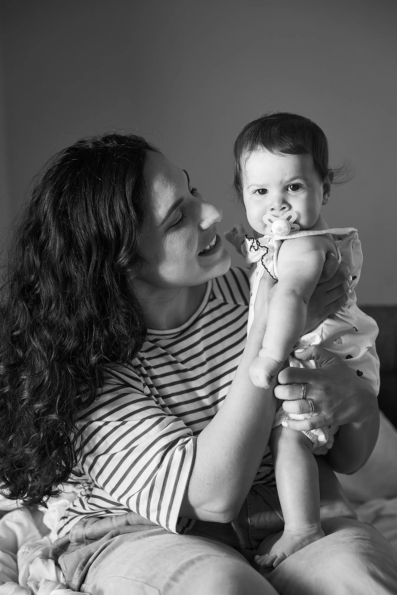 A woman with curly hair and a striped shirt holding a baby with a pacifier in her mouth, indoors, in a black and white photograph.