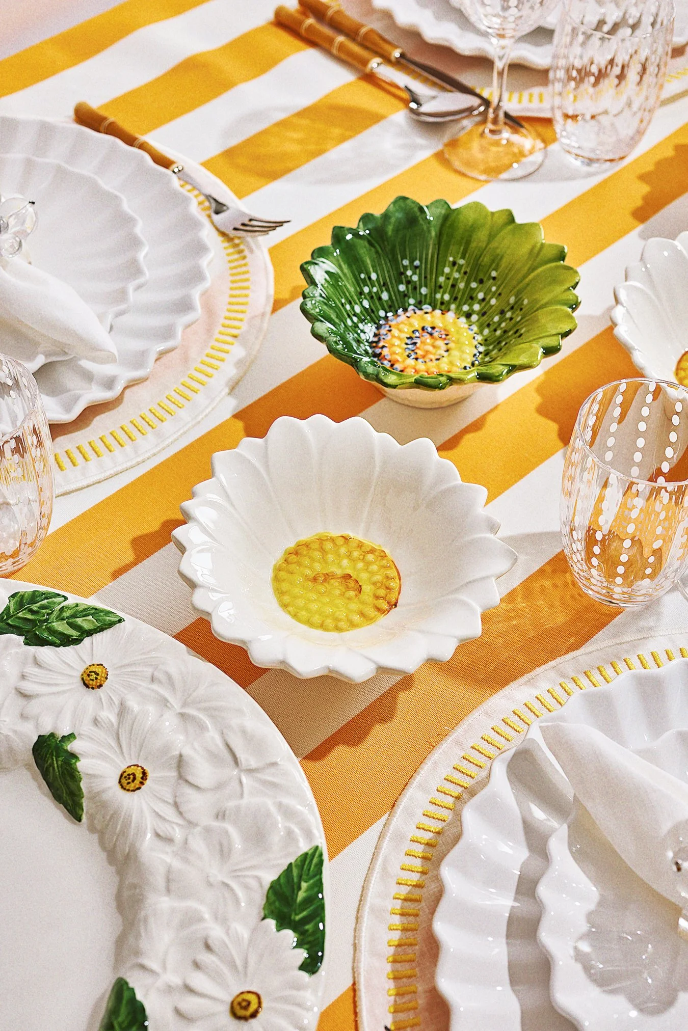 A table setting with yellow and white striped tablecloth, white decorative plates with floral and leaf designs, green and white ceramic bowls, clear glasses with dotted patterns, and silverware with bamboo-style handles.