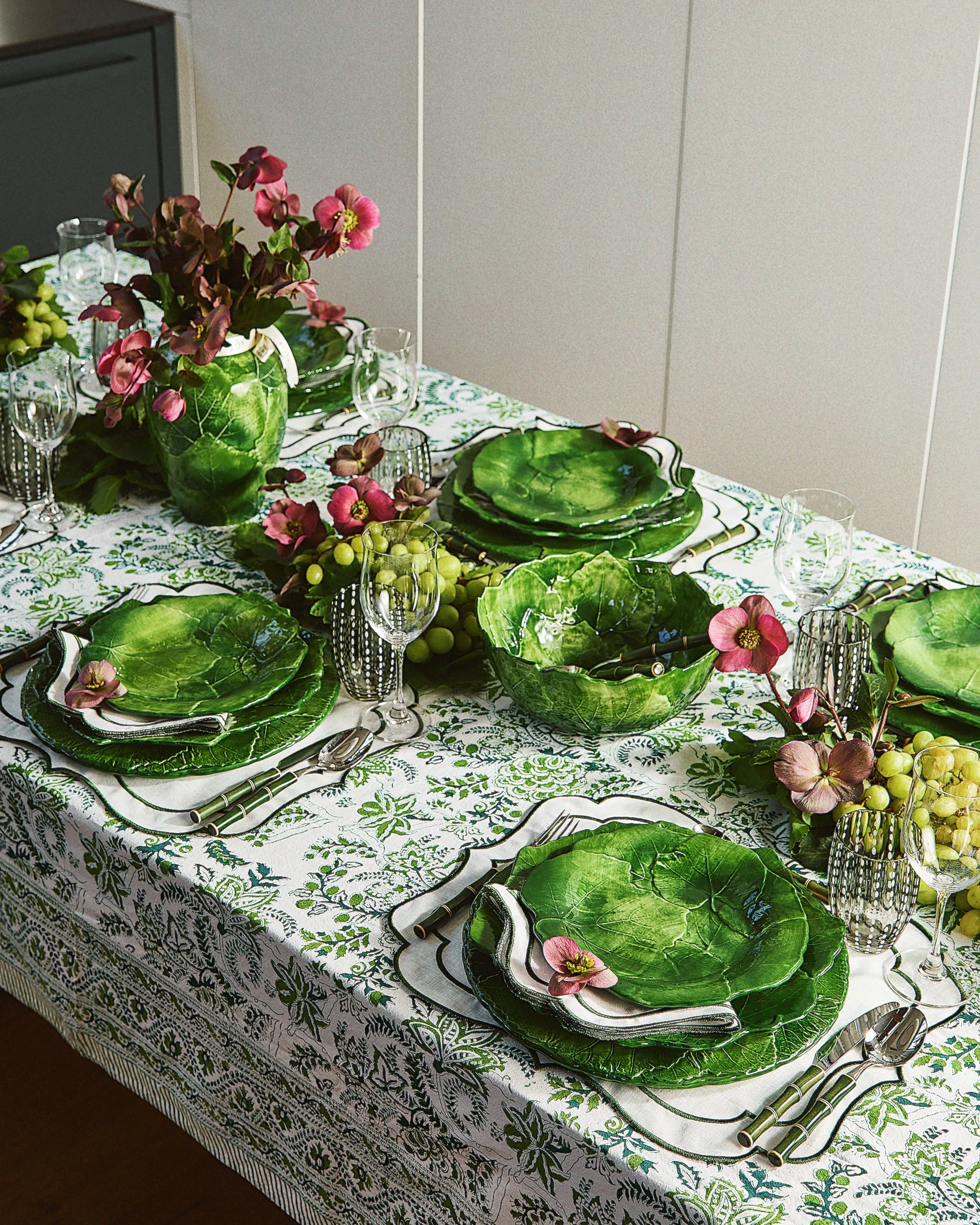 Table setting with green leaf-shaped plates, glasses, silverware, pink flowers, grapevine decorations, and a white tablecloth with green floral patterns.