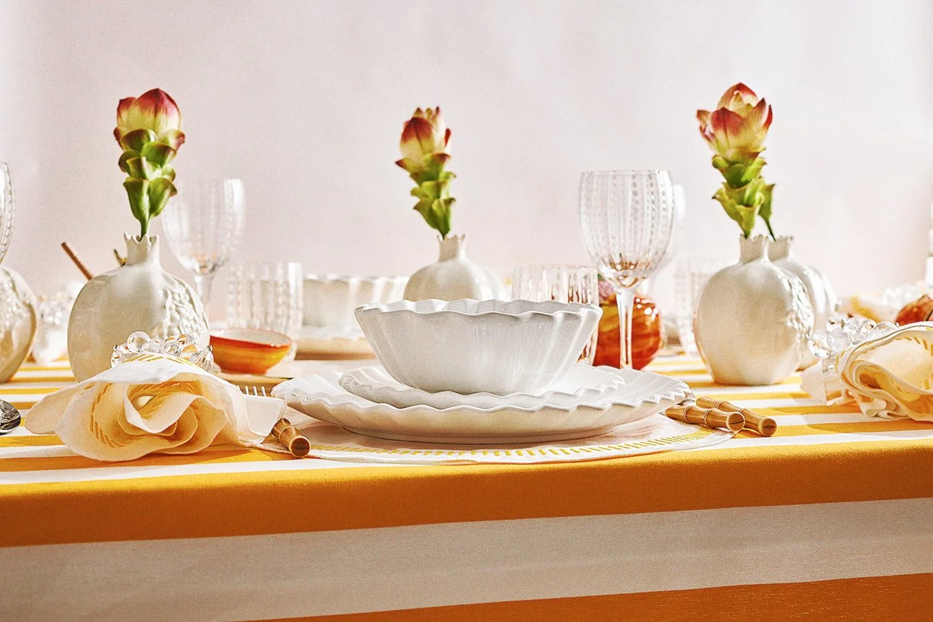 Table set with white ceramic bowls, clear glassware, yellow and white striped tablecloth, yellow napkins, and white vases with pink and green flowers.