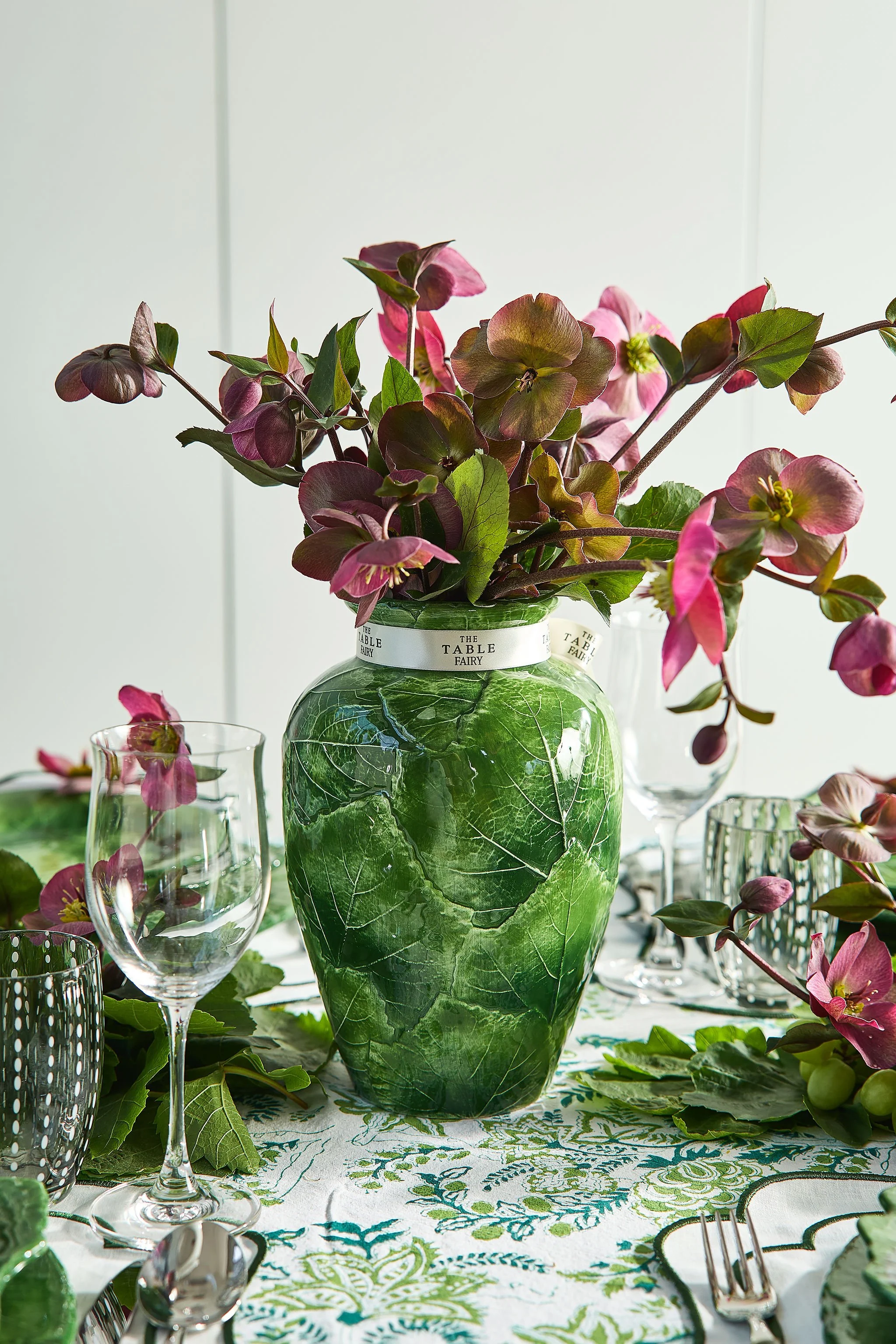 A table centerpiece featuring a large green leafy patterned ceramic vase with pink and purple flowers, surrounded by glasses and tableware, on a green and white embroidered tablecloth.