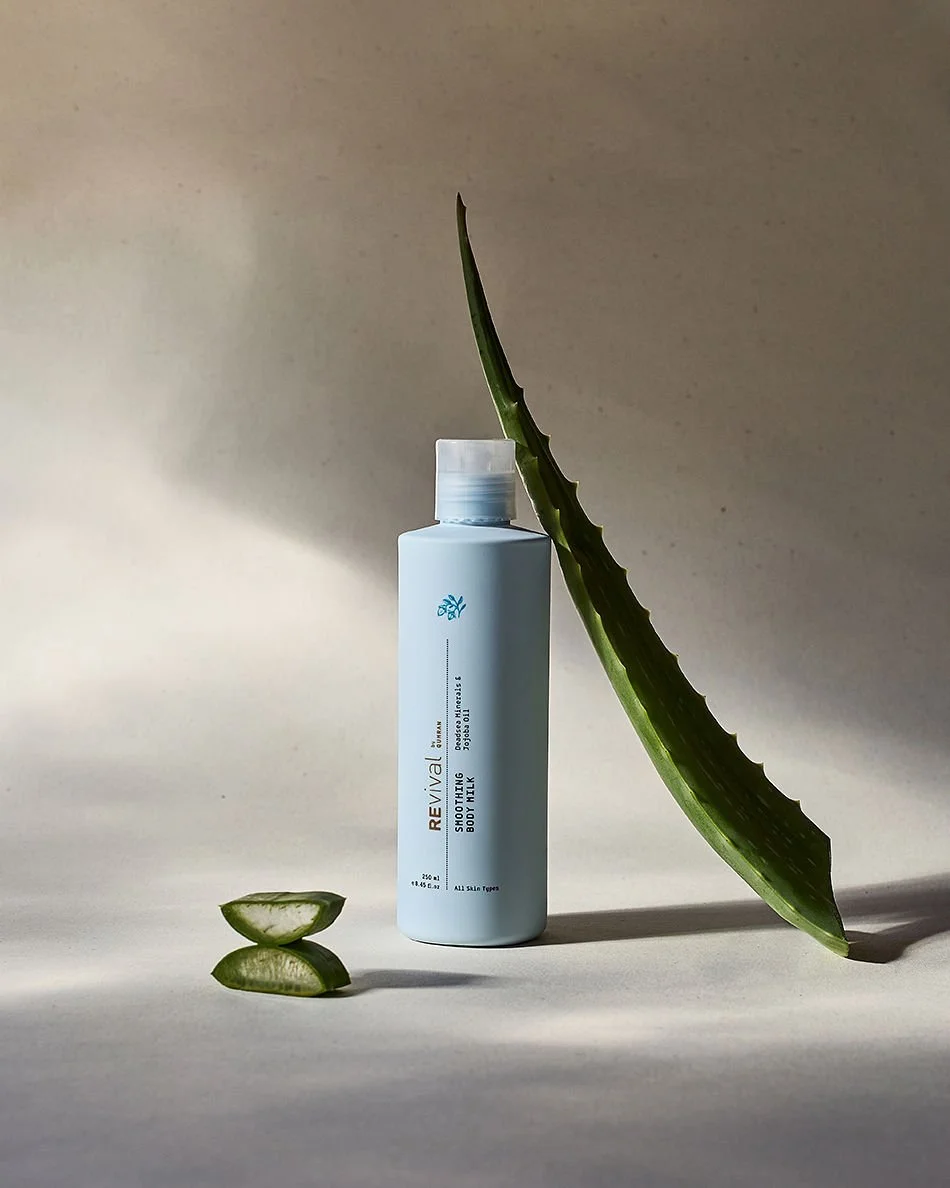 A white bottle of Revive skincare product with a large aloe vera leaf leaning against it, and a small aloe vera slice in front, on a light surface with a neutral background.