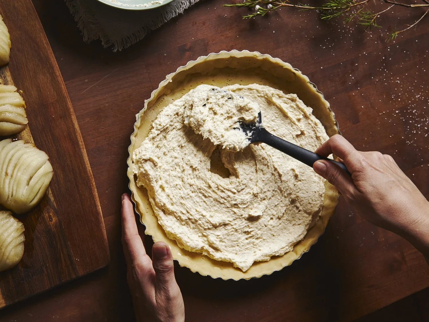 A person scooping filling into a pie crust with a black spatula, with apple slices on a wooden board to the side.