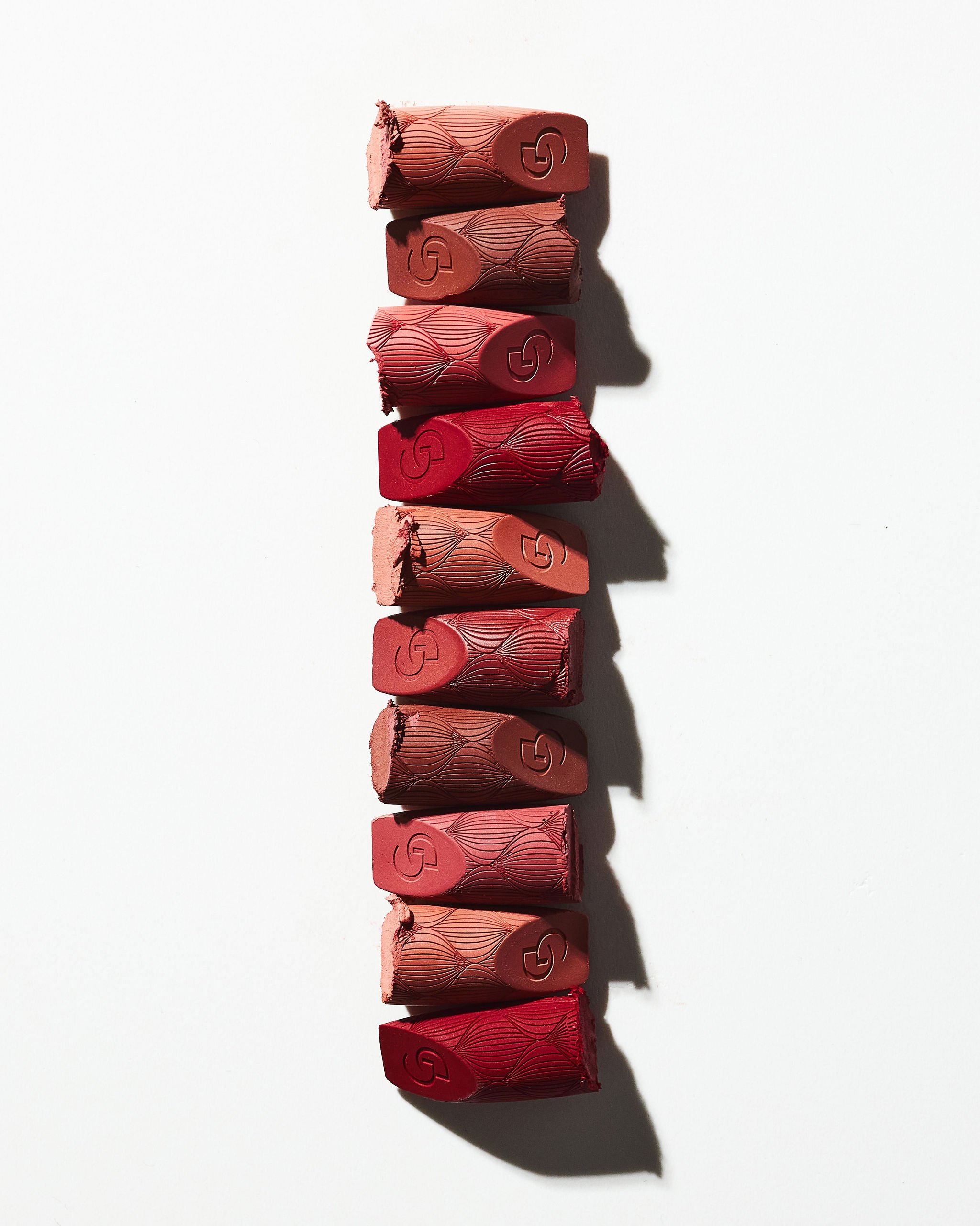 A row of ten torn lipstick bullets arranged vertically on a white background, showing various shades of red and pink with embossed designs.