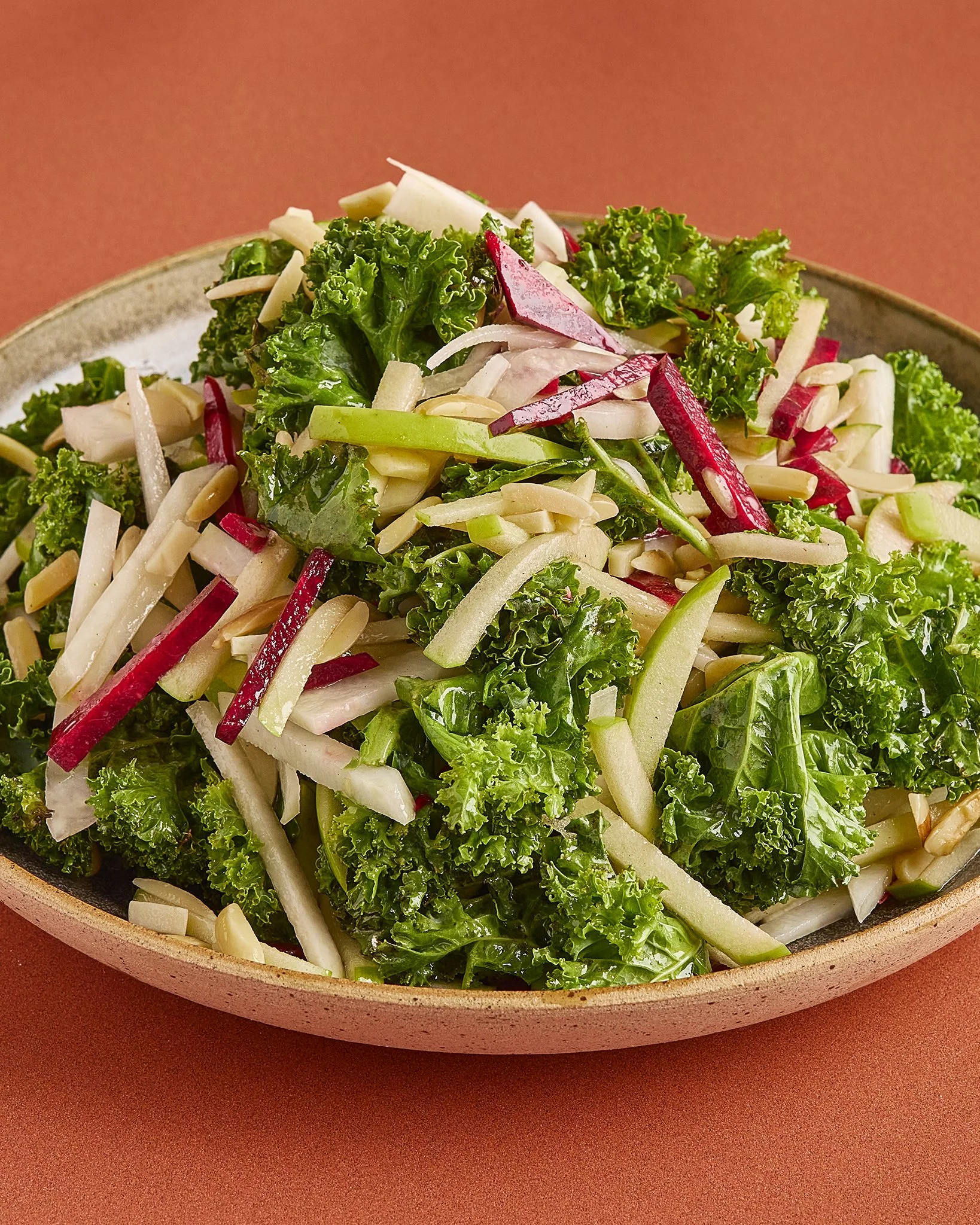 Bowl of fresh kale, radish, and cucumber salad with slivered almonds on a reddish-brown surface.